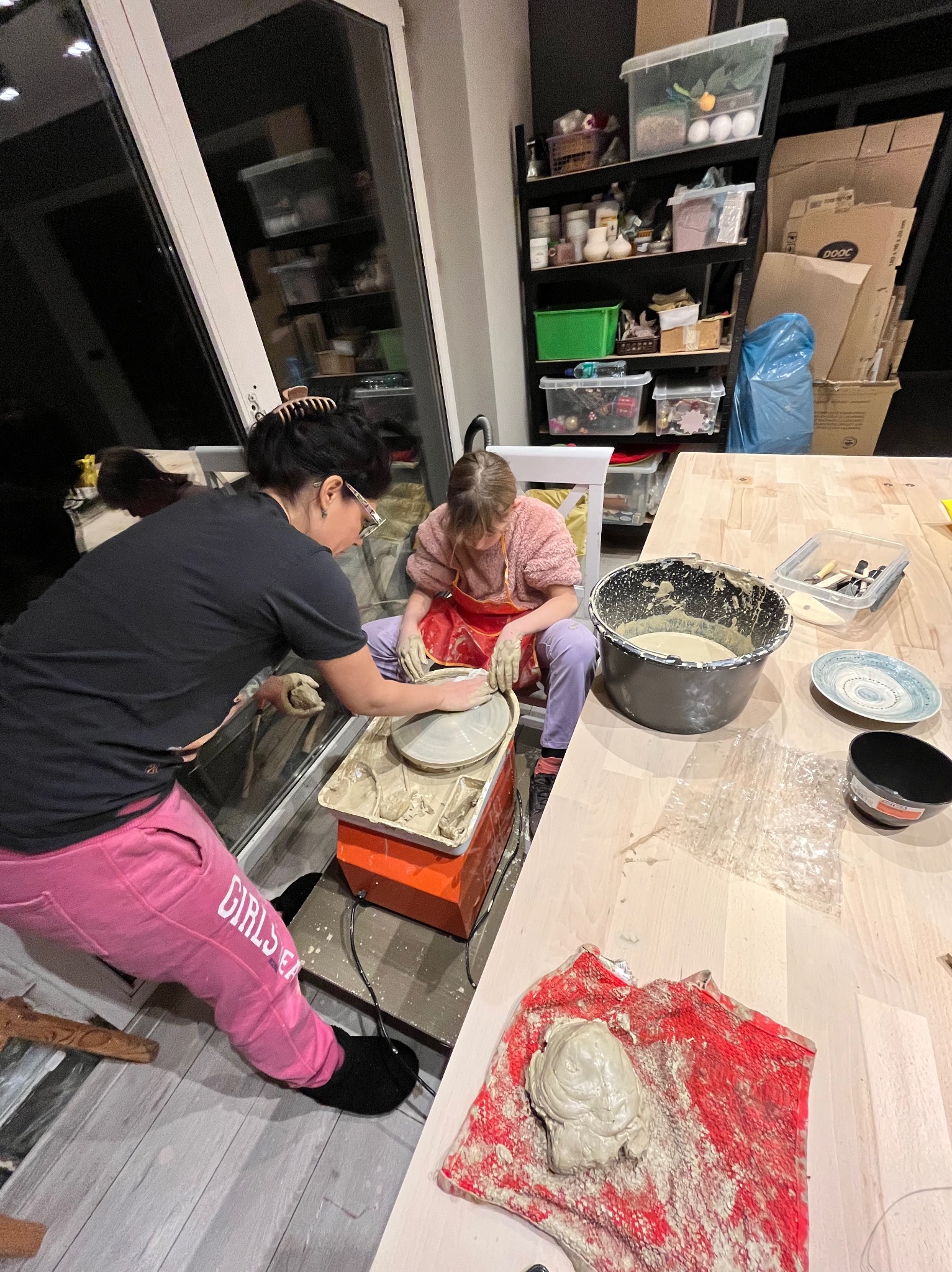 Kobieta z dziewczynką formują glinę na kole garncarskim w warsztacie, widoczny kubeł z gliną, narzędzia i gotowe wyroby.