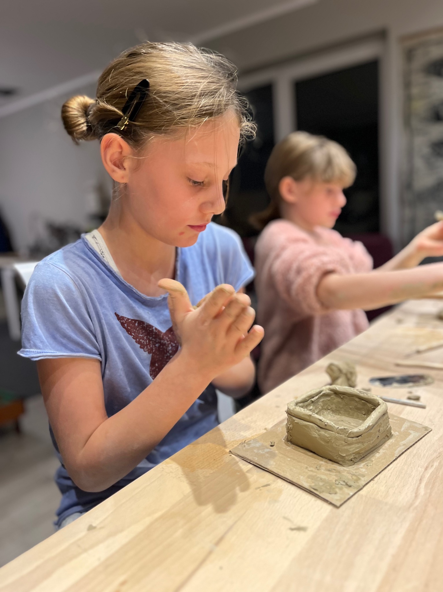 Dziewczynka formuje glinę przy drewnianym stole, tworząc małe naczynie; w tle druga dziewczynka również pracuje z gliną.