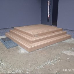 Rd systemy kompozytowe - Schody z desek przy szarym budynku. Trzy stopnie prowadzą do drzwi. Widoczna krata ściekowa i ziemia. Jasne, naturalne światło.