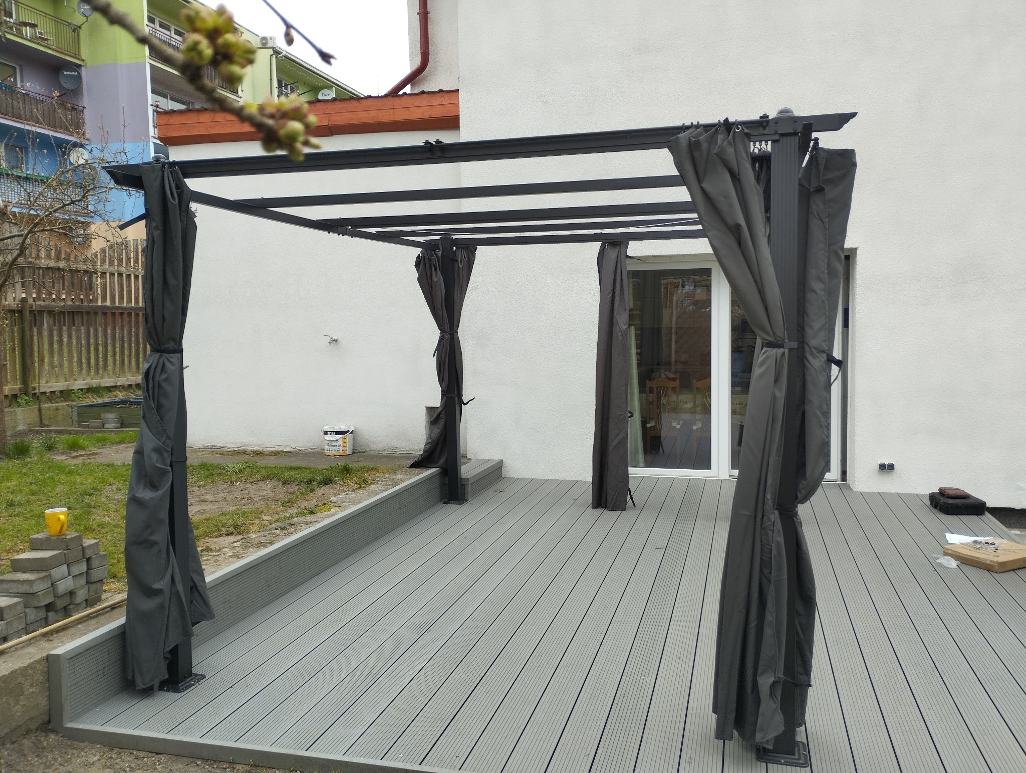Szara, nowoczesna pergola z zasłonami na tarasie z desek. Widoczny fragment domu z białymi ścianami. Na tarasie leżą narzędzia i akcesoria montażowe.