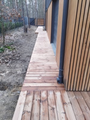 Długa, drewniana ścieżka z desek prowadząca wzdłuż fasady budynku z drewnianą okładziną. Widoczne drzewa w tle i czarna rura spustowa.