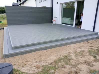 Nowoczesny taras z szarych desek z dwoma stopniami, otoczony piaskiem, z minimalistycznym krzesłem i panelem osłonowym w tle. Osoba w drzwiach przesuwnych.