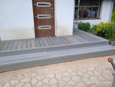 Szary taras kompozytowy z dwoma stopniami prowadzący do brązowych drzwi wejściowych domu. Widok na fragment ogrodu z oknem w tle.