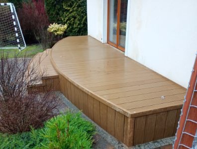 Nowoczesny taras z brązowych desek, z zaokrąglonym wykończeniem, prowadzący do drzwi balkonowych. Widoczne schody i elementy zieleni. W tle siatka do piłki nożnej.