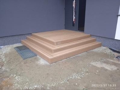 Schody z desek przy szarym budynku. Trzy stopnie prowadzą do drzwi. Widoczna krata ściekowa i ziemia. Jasne, naturalne światło.