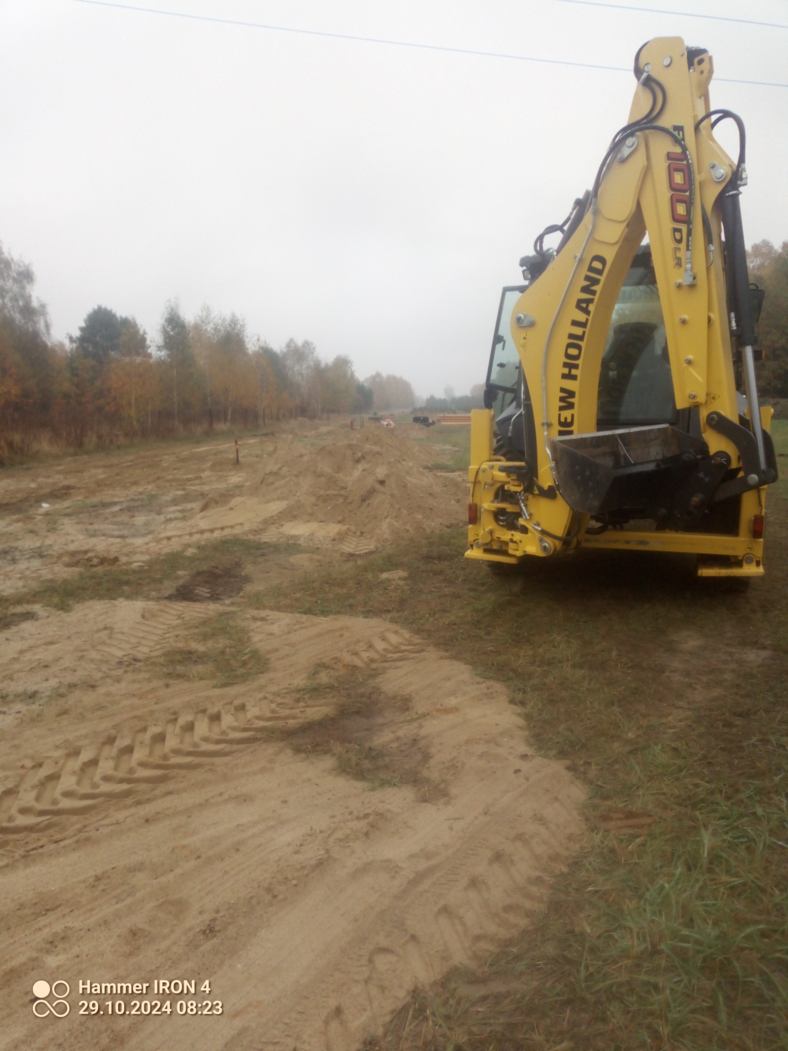Żółta koparko-ładowarka New Holland na terenie budowy, widoczne świeże ślady opon na piasku i usypana ziemia w tle, pochmurny dzień.