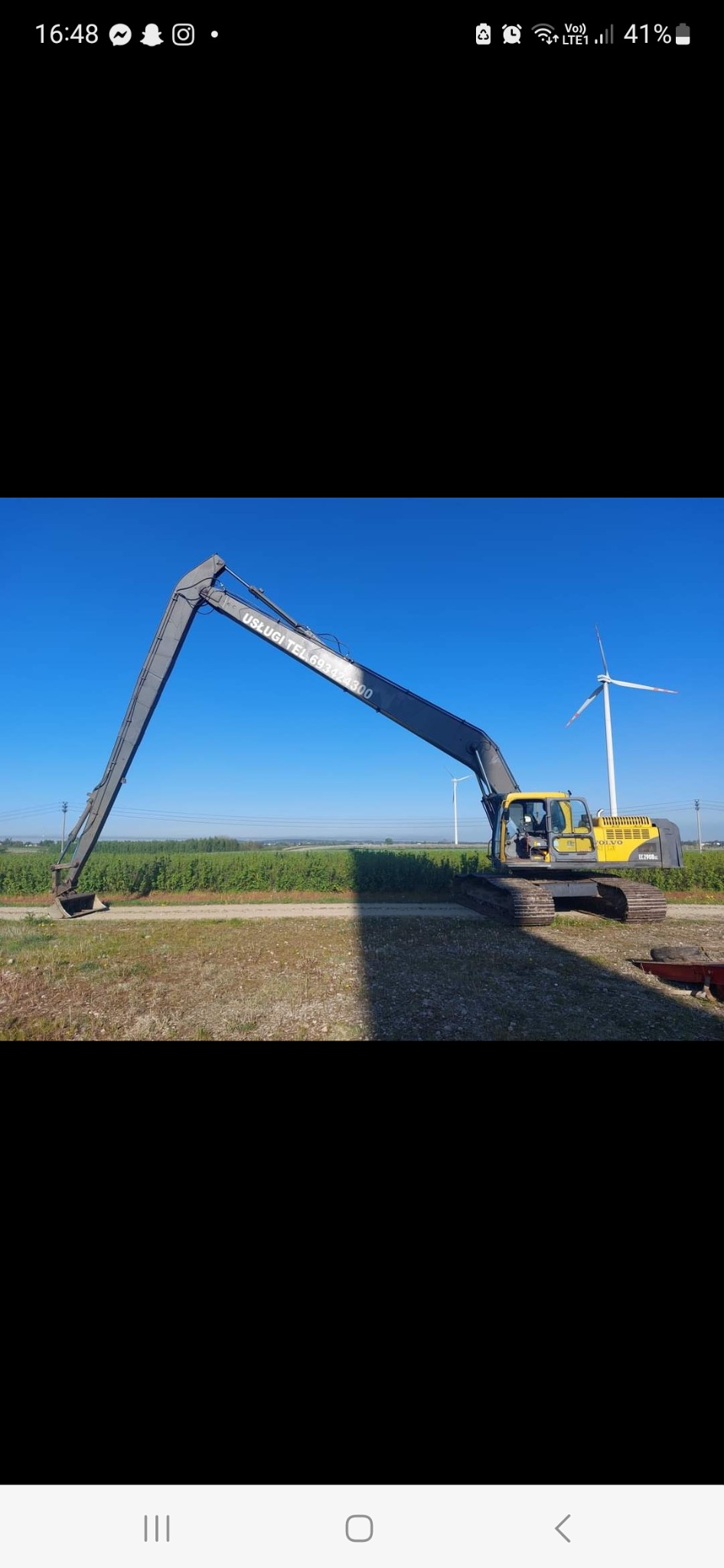 Żółta koparka Volvo EC290BLC z długim ramieniem na tle pola i turbin wiatrowych, z widocznym numerem telefonu na ramieniu