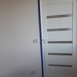 MANS-BUD - Białe drzwi wewnętrzne z poziomymi, szarymi paskami, osadzone w ościeżnicy zabezpieczonej niebieską taśmą malarską, widoczne gniazdka elektryczne na ścianie i kable przy podłodze.