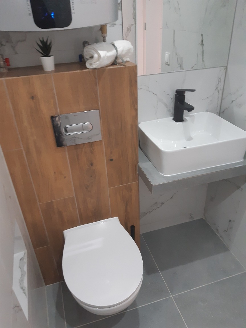 Nowoczesna łazienka z wiszącą miską WC, umywalką nablatową z czarną armaturą, drewnopodobną zabudową i marmurowymi płytkami na ścianach.