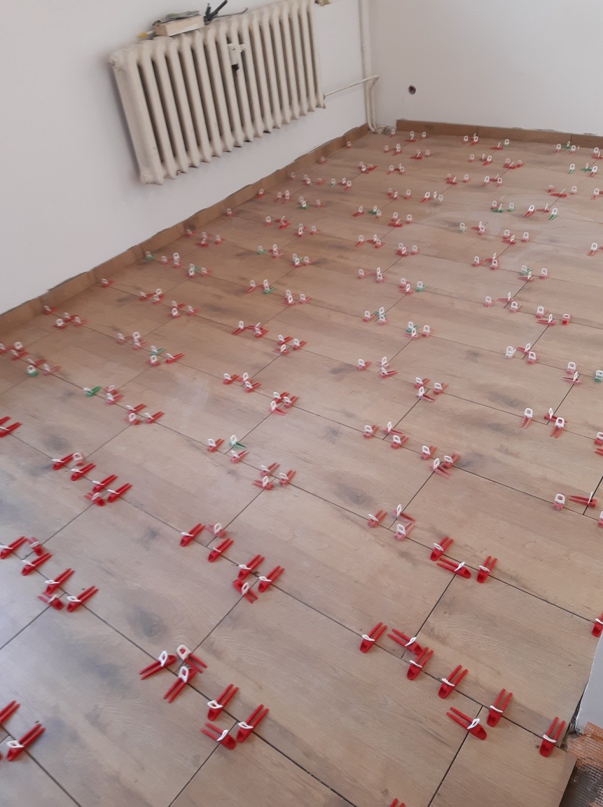 Ułożone płytki podłogowe imitujące drewno, z czerwonymi i zielonymi klipsami poziomującymi, widoczny fragment grzejnika żeliwnego przy ścianie.