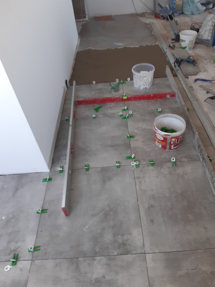 Układanie szarych płytek podłogowych z użyciem poziomicy i klipsów dystansowych, widoczne wiaderka z klejem i akcesoriami na częściowo przygotowanym podłożu.