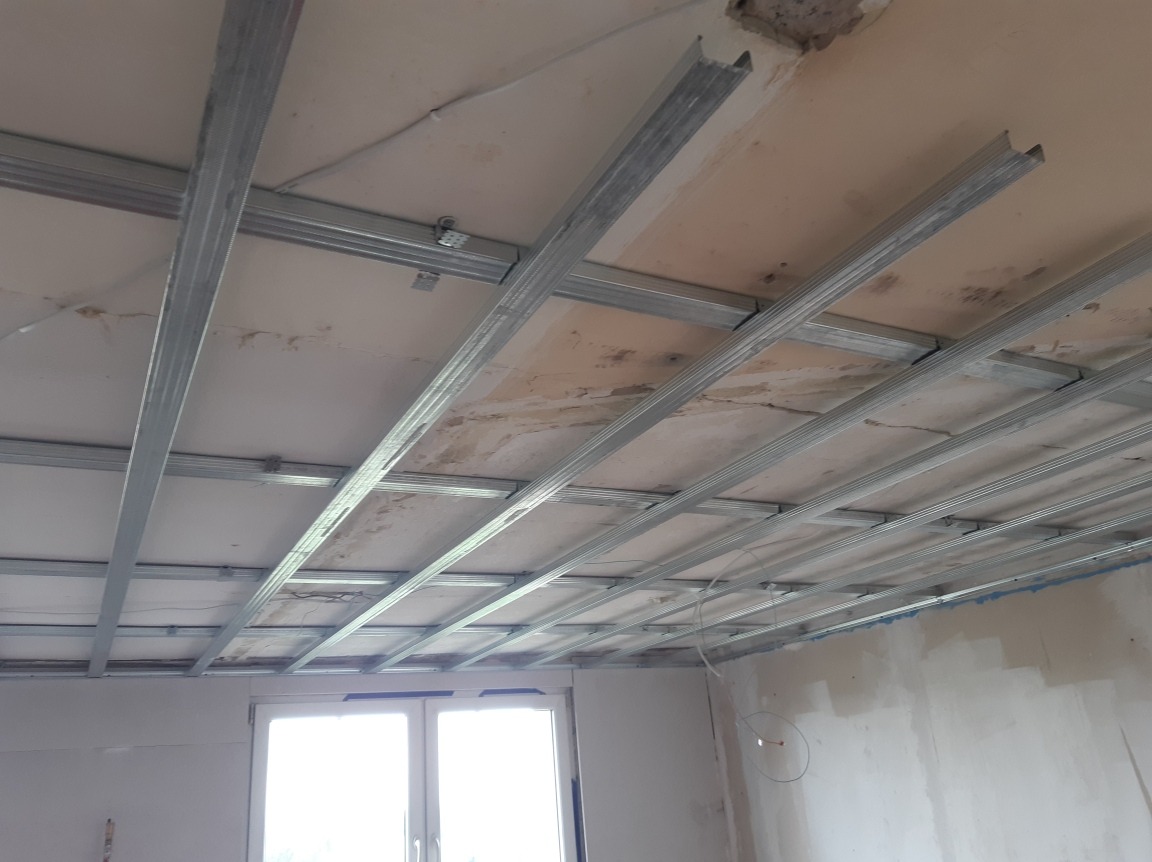 Metalowa konstrukcja sufitu podwieszanego zamocowana do starego sufitu w trakcie prac wykończeniowych, widoczne profile i elementy montażowe, okno w tle.