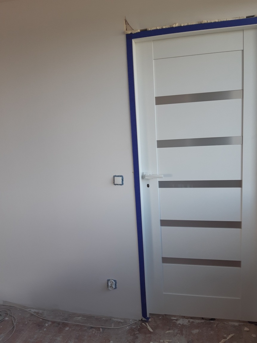 Białe drzwi wewnętrzne z poziomymi, szarymi paskami, osadzone w ościeżnicy zabezpieczonej niebieską taśmą malarską, widoczne gniazdka elektryczne na ścianie i kable przy podłodze.