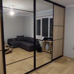 Piotr Andrukh - Duża szafa z przesuwnymi drzwiami lustrzanymi i drewnianymi panelami, czarna rama, widok na wnętrze pokoju z szarą sofą narożną i parkietem w jodełkę.