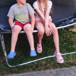 Piotr Andrukh - Dwoje dzieci siedzi na trampolinie ogrodowej z siatką ochronną, na tle zieleni.