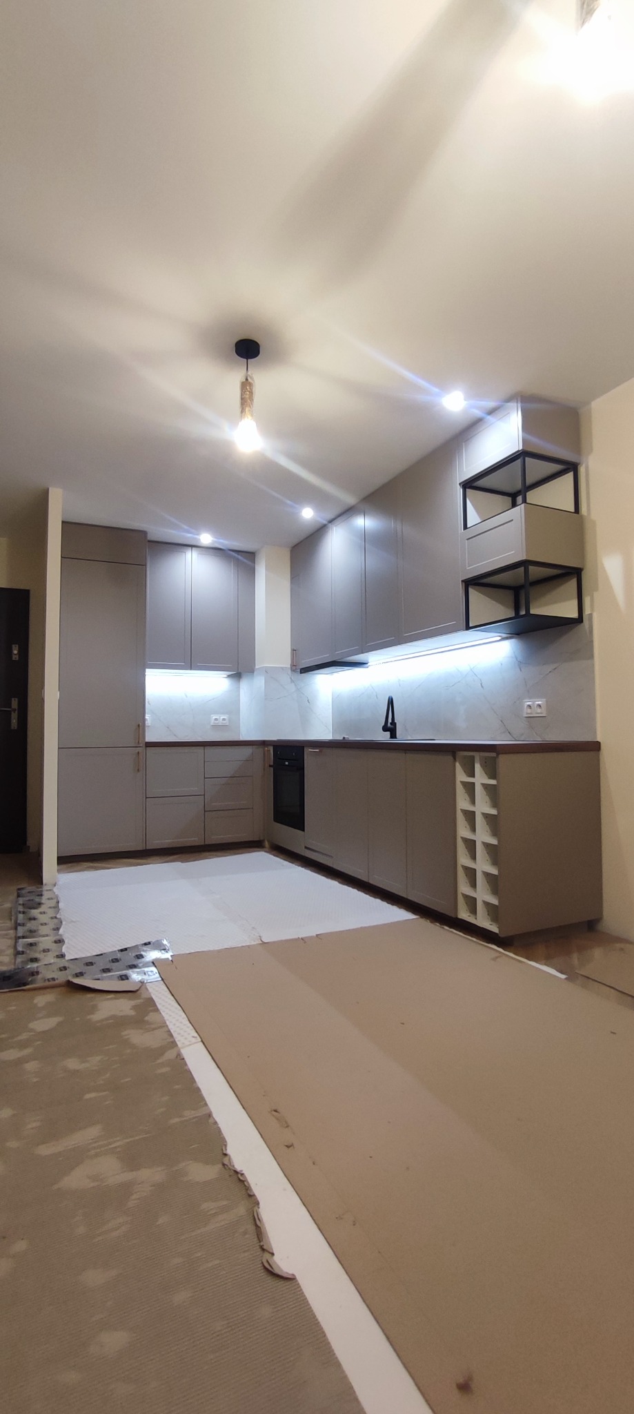 Jasnoszara kuchnia na wymiar w trakcie montażu, widoczne zabezpieczenia podłogi, oświetlenie podszafkowe LED, marmurowy backsplash i czarna armatura.