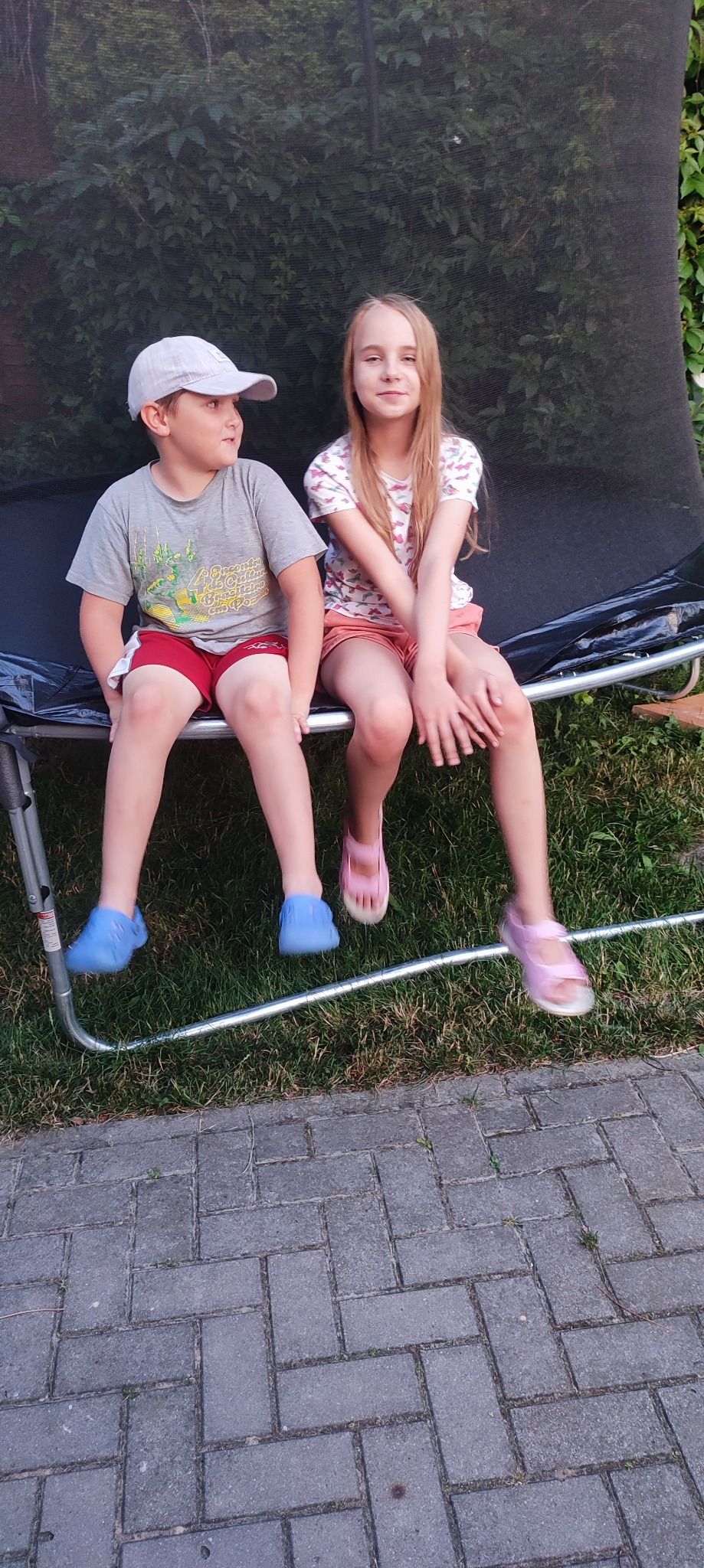 Dwoje dzieci siedzi na trampolinie ogrodowej z siatką ochronną, na tle zieleni.
