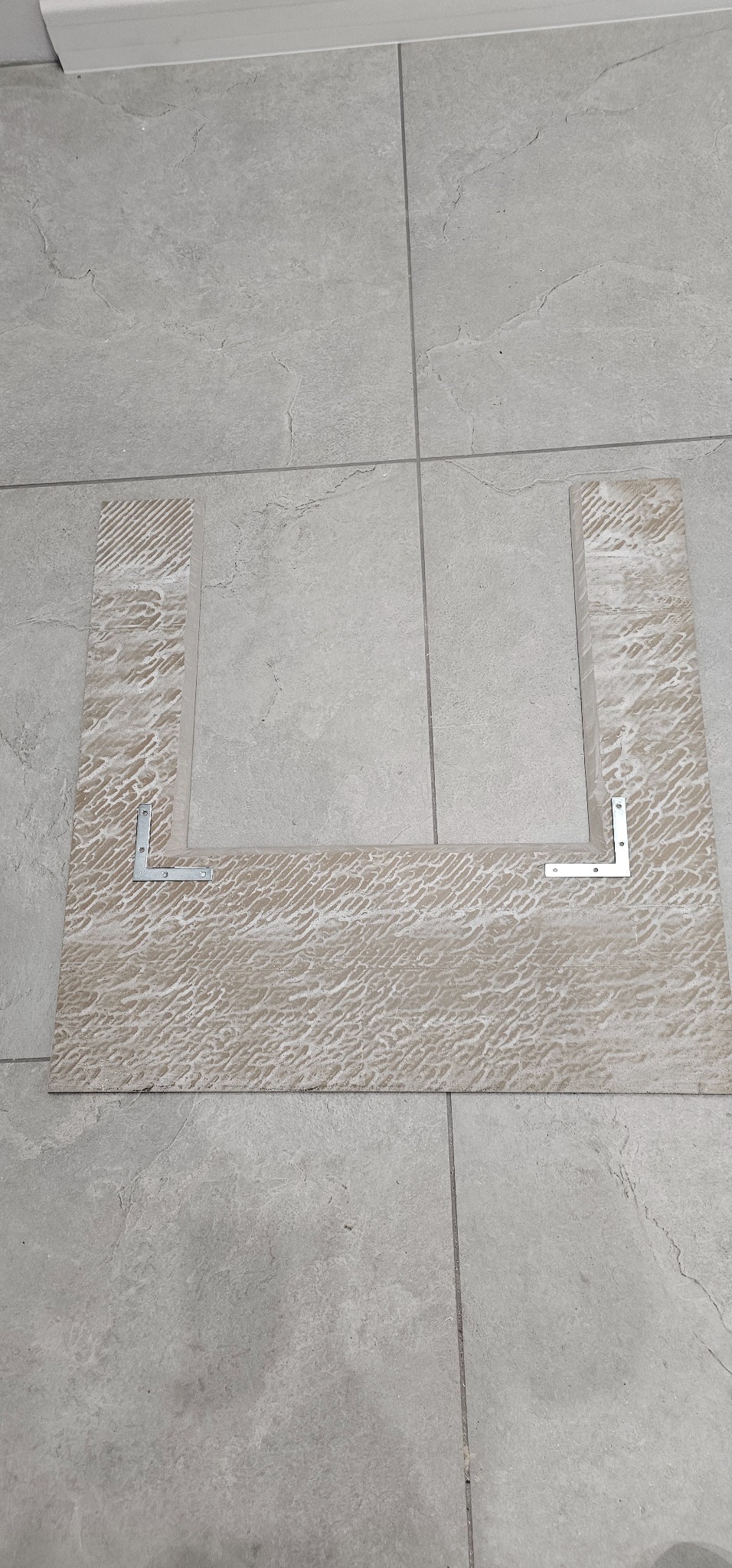 Wycięty fragment beżowej płytki podłogowej z wzorem, ułożony na szarych płytkach, z przymocowanymi metalowymi kątownikami.