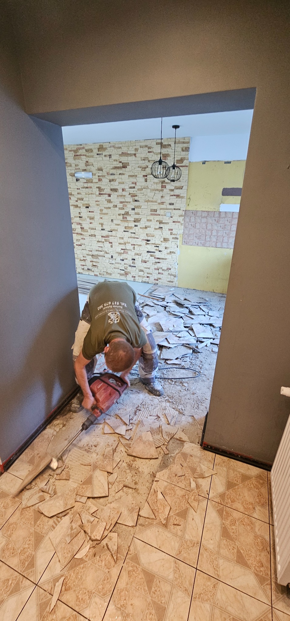 Pracownik w trakcie skuwania starych, beżowych płytek podłogowych za pomocą młota udarowego; widoczne fragmenty skruszonych płytek i przygotowane ściany do dalszych prac.