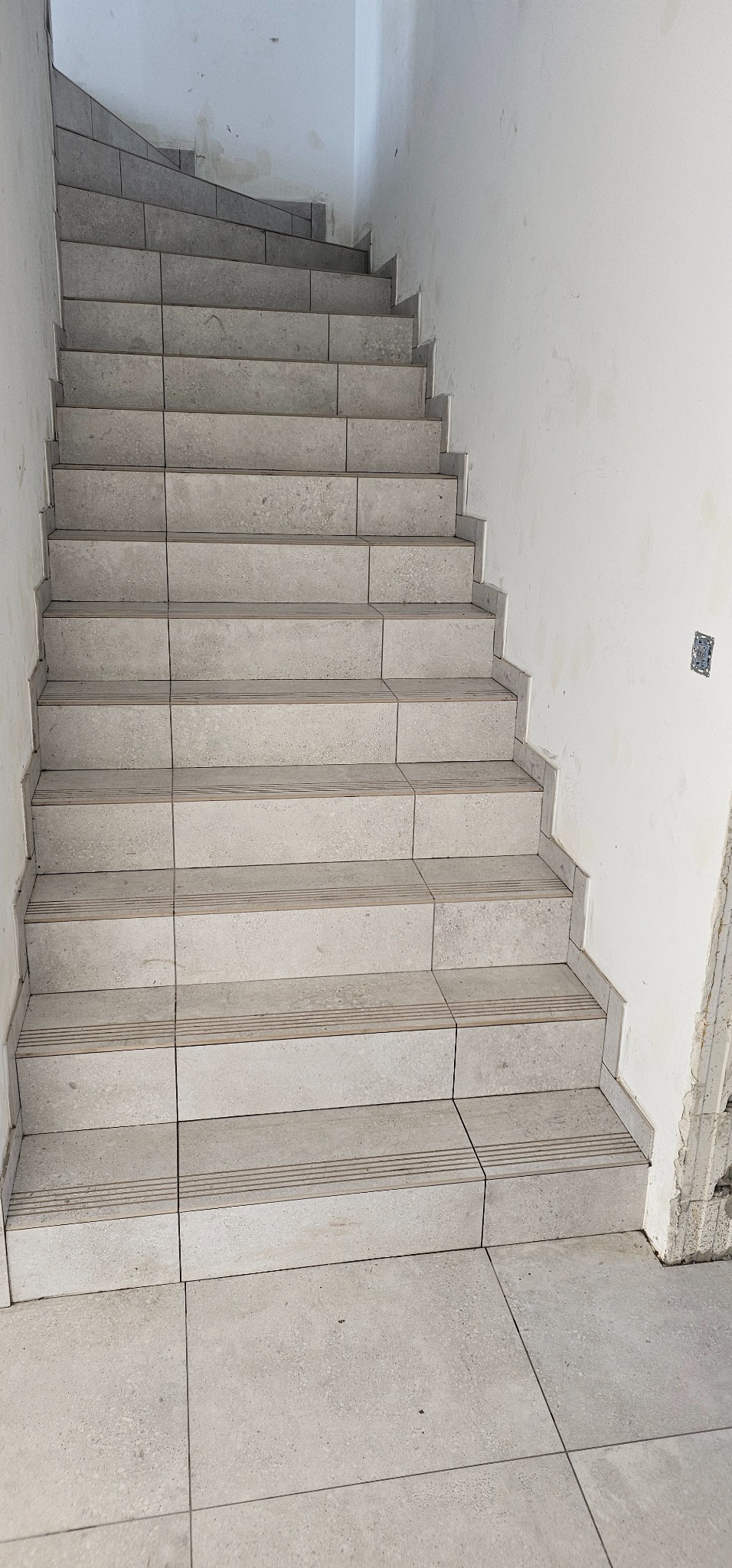 Schody wyłożone jasnoszarymi płytkami ceramicznymi, widok z dołu na górę, ściany w stanie surowym, widoczne niedoskonałości i ślady po pracach budowlanych.