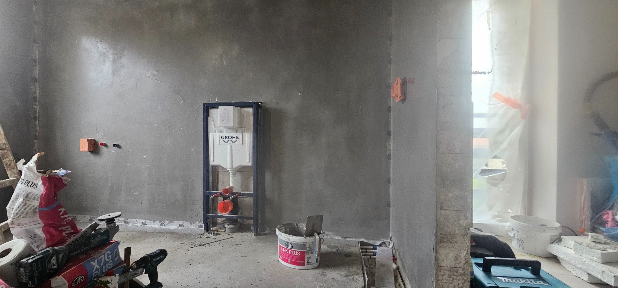 Wnętrze w trakcie prac wykończeniowych: ściany po nałożeniu szpachli, widoczny stelaż podtynkowy WC marki Grohe, narzędzia budowlane i worki z zaprawą Ardex S1-K Plus na podłodze, zabezpieczone...