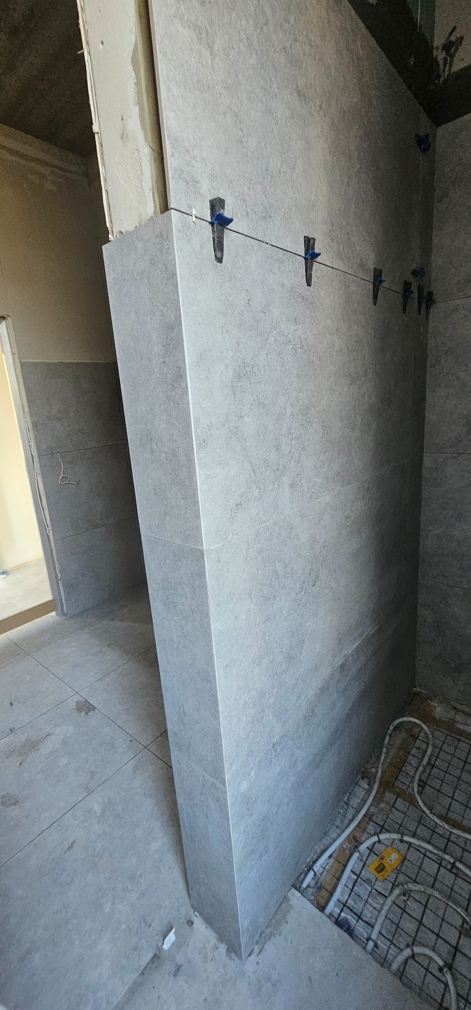 Montaż szarej, matowej płytki ściennej z użyciem klipsów poziomujących; widoczne ogrzewanie podłogowe w tle.