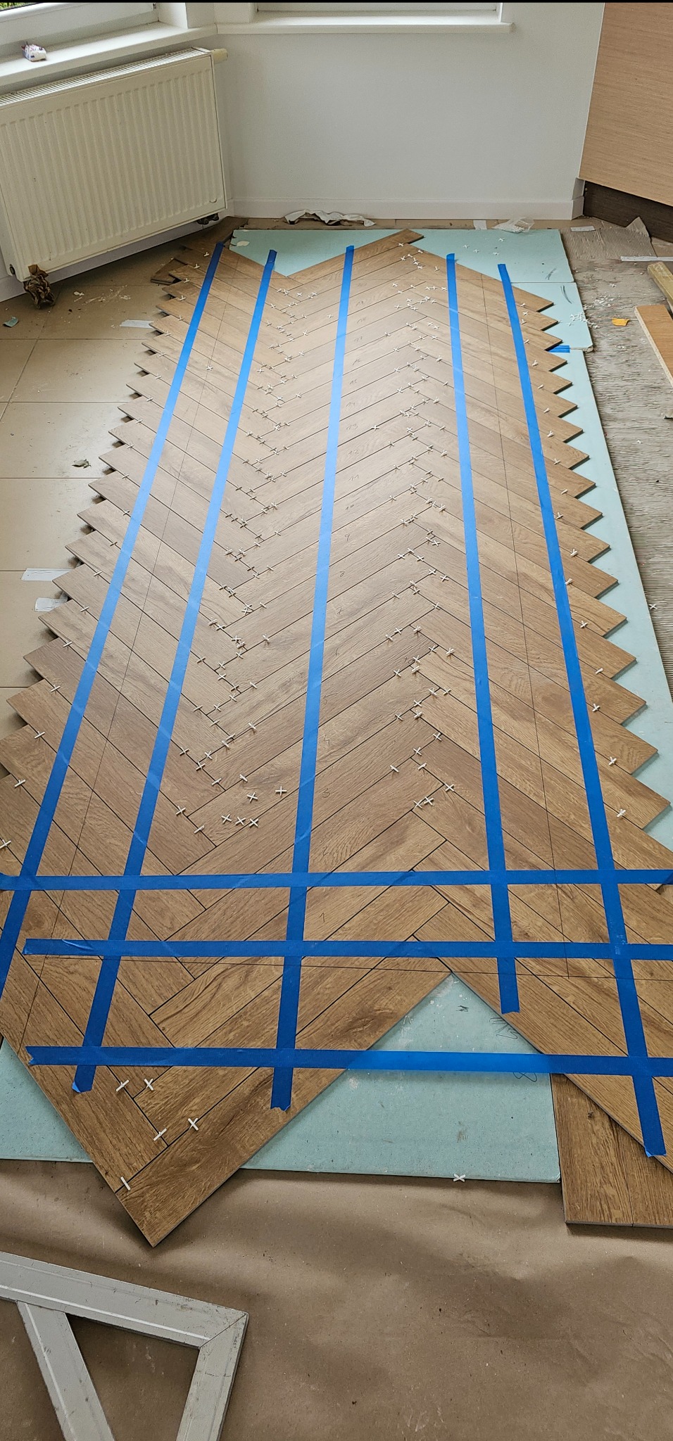 Układanie parkietu w jodełkę z użyciem niebieskiej taśmy maskującej i krzyżyków dystansowych, widoczne niedocięte panele i podkład.