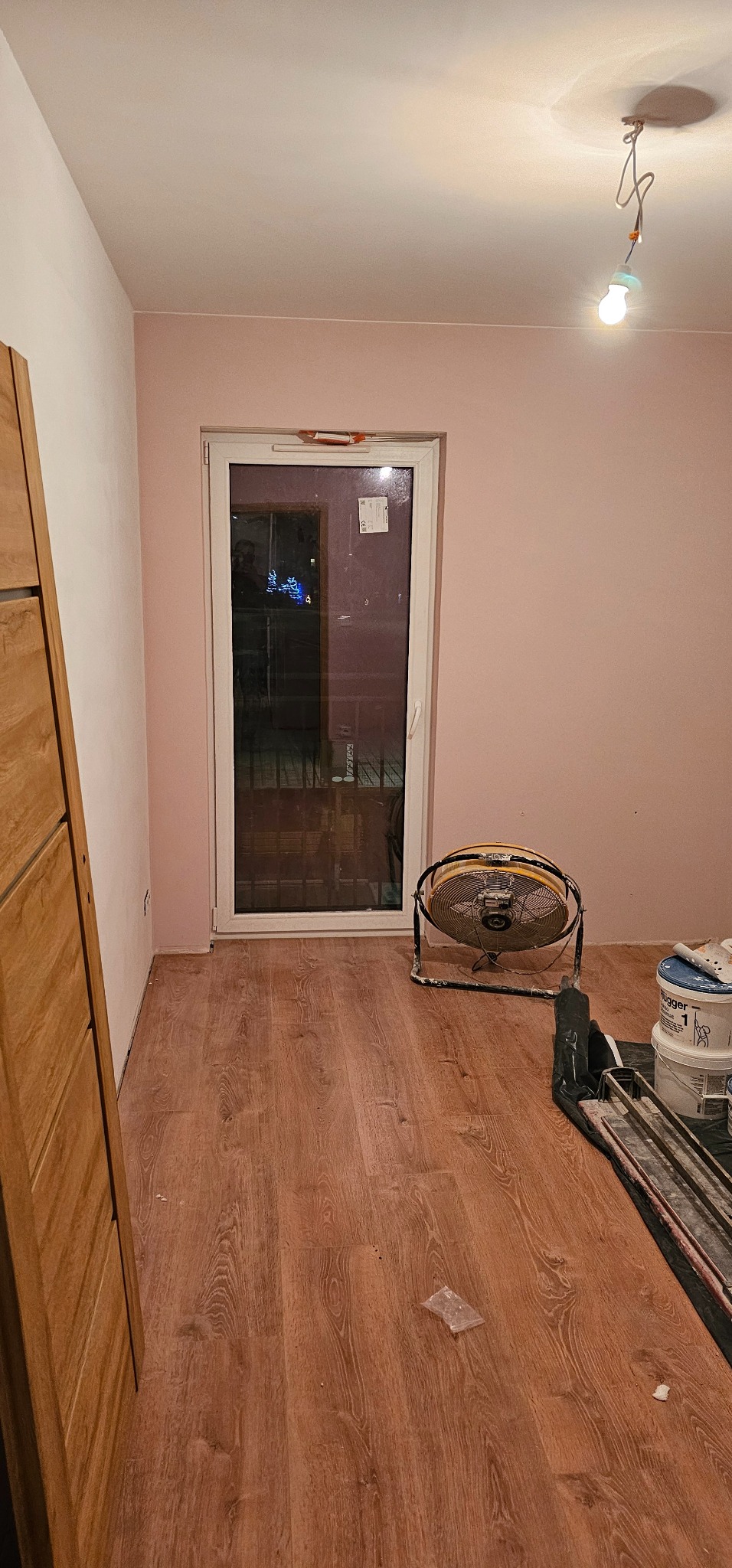 Pomieszczenie w trakcie prac wykończeniowych: różowe ściany, drewnopodobne panele podłogowe, wentylator pod oknem, wiaderka z farbą i narzędzia glazurnicze w rogu.
