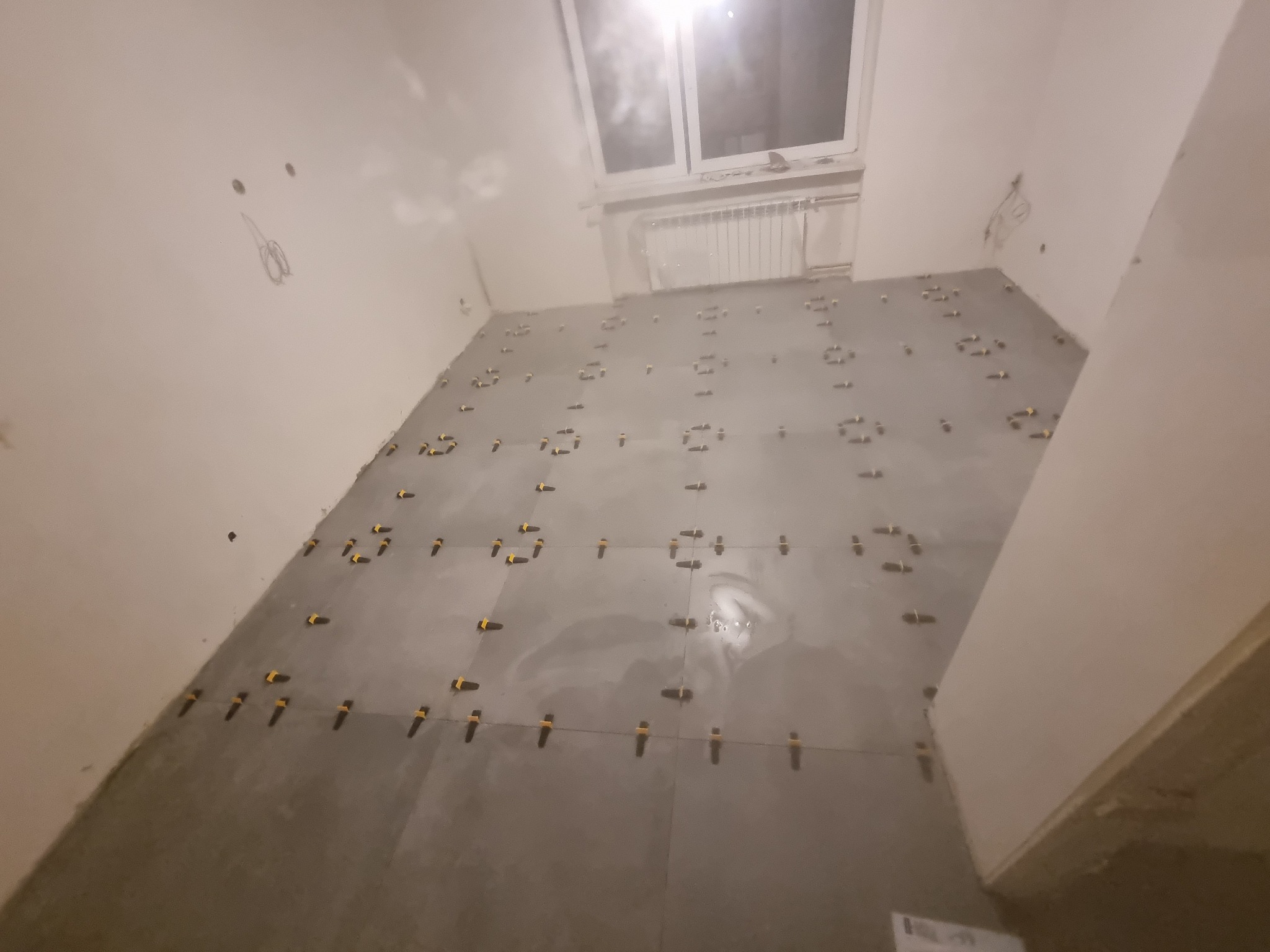 Układanie szarych płytek podłogowych w pomieszczeniu ze skośnym oknem dachowym, widoczne żółte klipsy poziomujące płytki, ściany przygotowane do dalszych prac wykończeniowych.