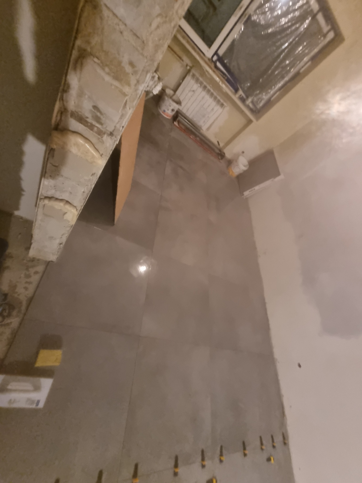 Szare płytki podłogowe, świeżo ułożone, z widocznymi krzyżykami dystansowymi, fragment okna zabezpieczonego folią oraz niedokończone obramowanie sufitu.