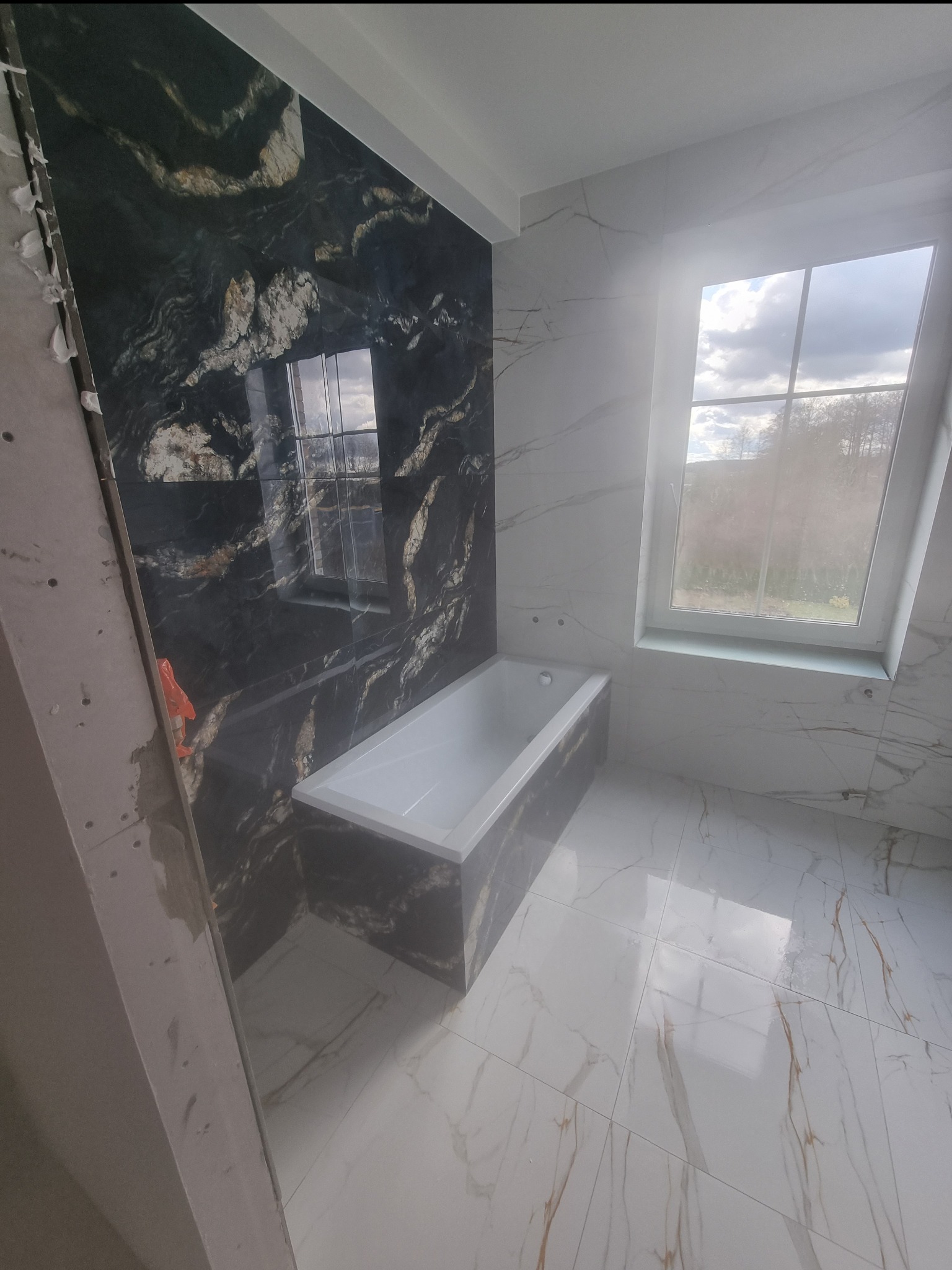 Wnętrze łazienki w trakcie prac wykończeniowych, widoczna wanna obudowana czarnym marmurem, białe marmurowe płytki na podłodze i ścianie z oknem, fragment ściany z płytą gipsową w surowym stanie.