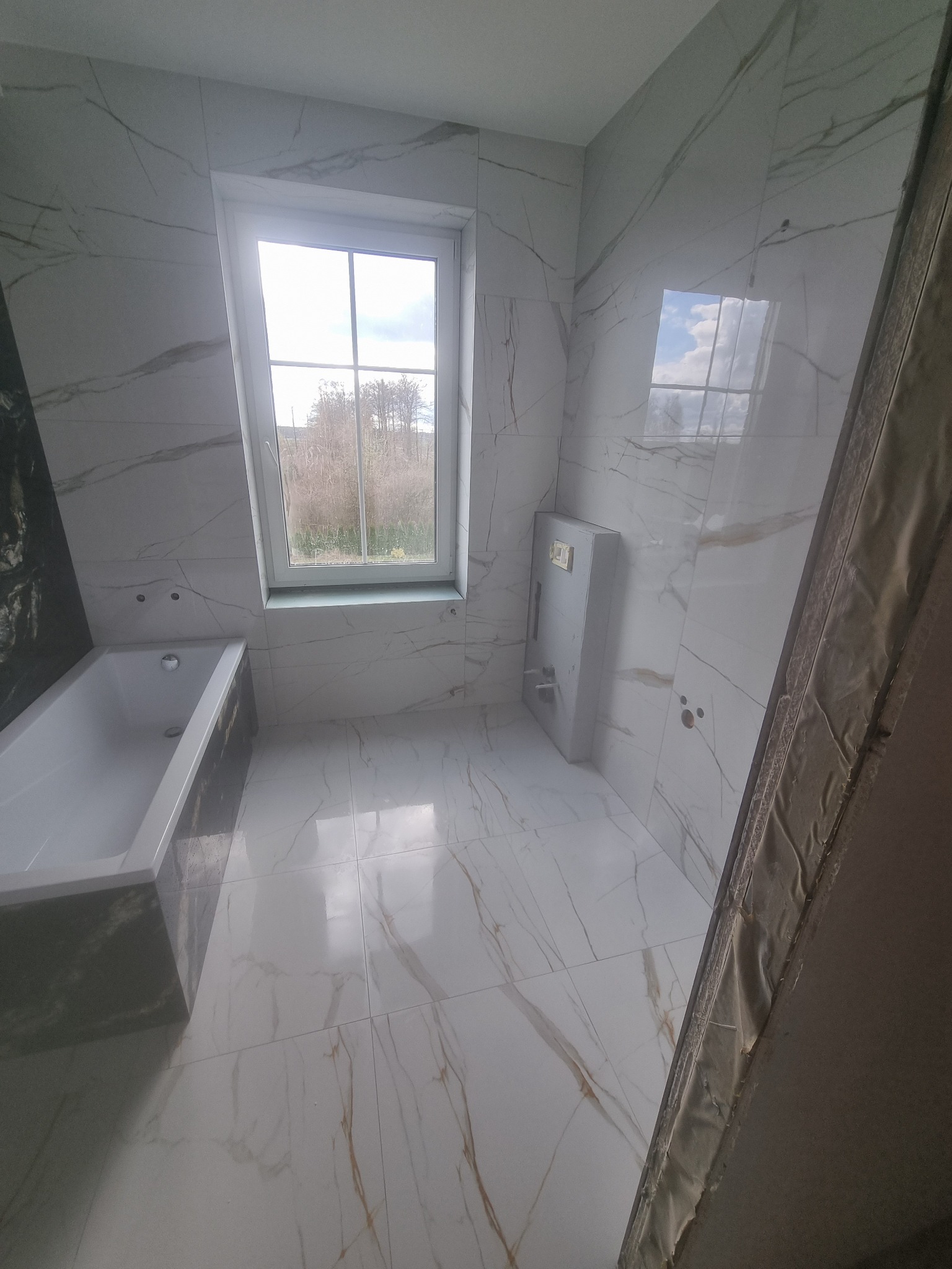 Nowoczesna łazienka z biało-złotymi płytkami imitującymi marmur, z widoczną wanną, oknem z widokiem na drzewa i zabudowaną spłuczką WC