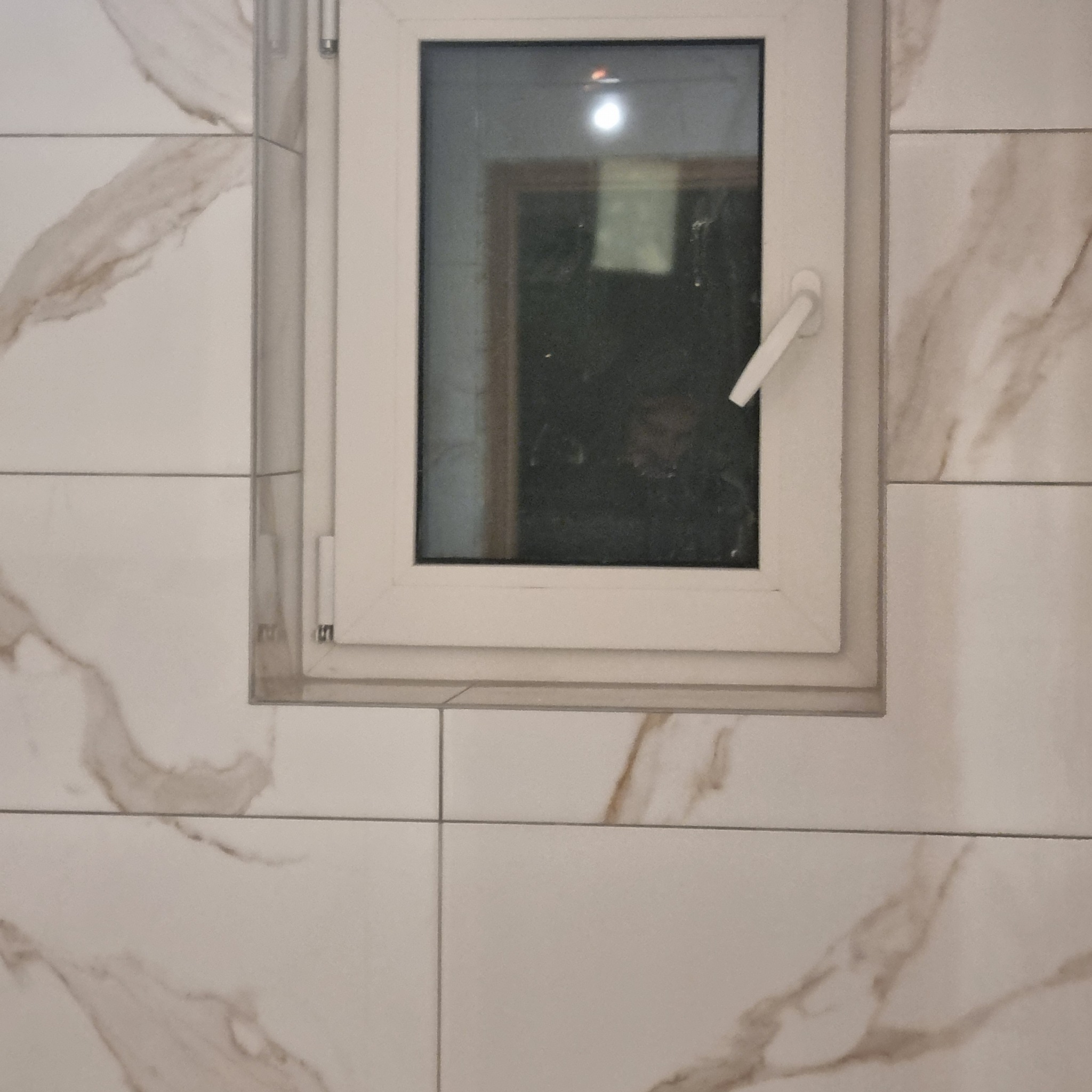 Ściana wyłożona białymi płytkami imitującymi marmur z wbudowanym białym oknem PCV z klamką