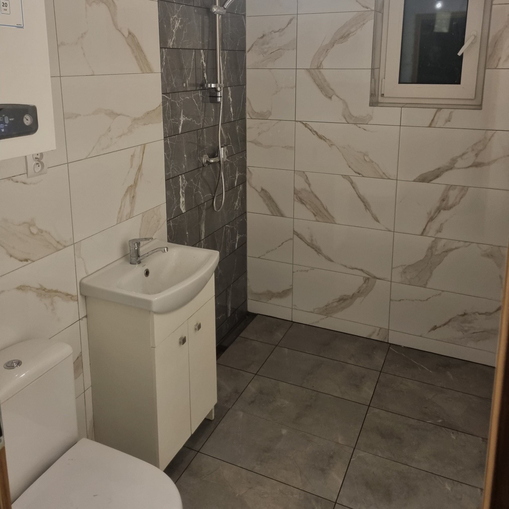 Wykończona łazienka z białymi i szarymi marmurkowymi płytkami na ścianach, szarą podłogą, białą szafką pod umywalką i toaletą w dolnym rogu.