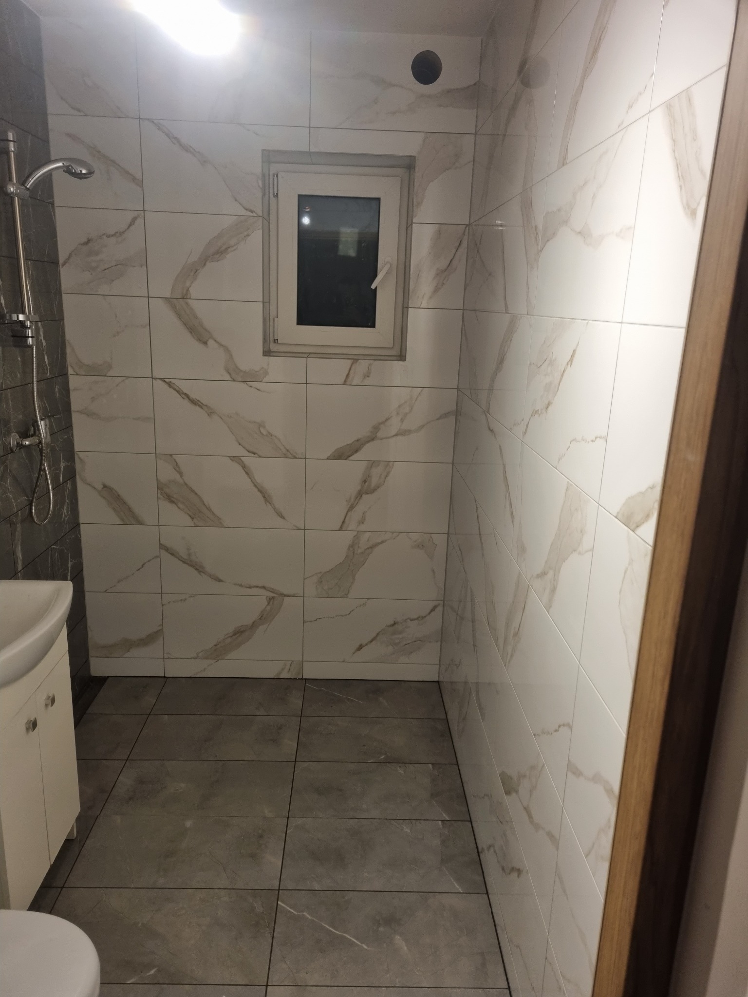 Wykończona łazienka z biało-szarymi marmurkowymi płytkami na ścianach i szarymi płytkami podłogowymi, widoczny fragment umywalki z szafką, kabina prysznicowa i okno