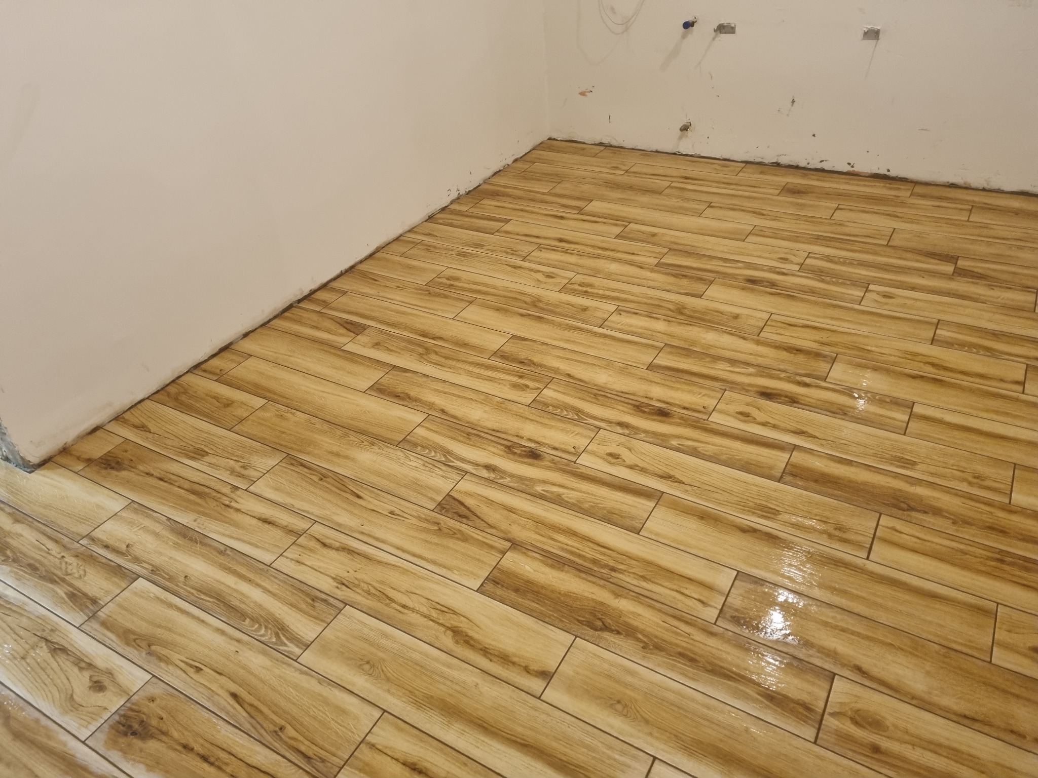 Świeżo ułożona, błyszcząca podłoga z płytek imitujących drewno w odcieniach beżu i brązu, ułożonych w jodełkę, widok na fragment ściany z widocznymi instalacjami.