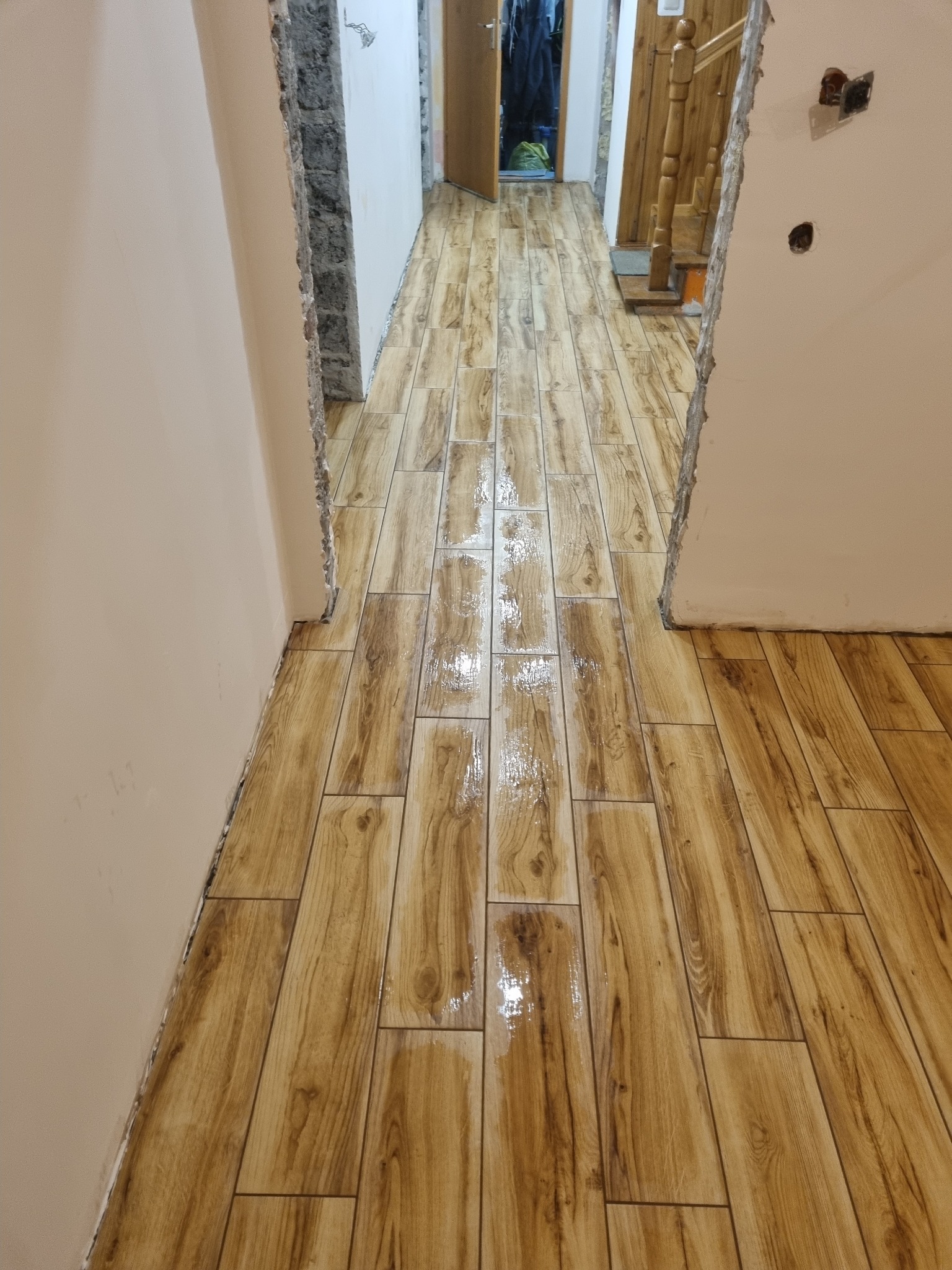 Korytarz w trakcie remontu z nową, błyszczącą podłogą z płytek imitujących drewno, widoczne niedokończone ściany i wystające kable elektryczne.