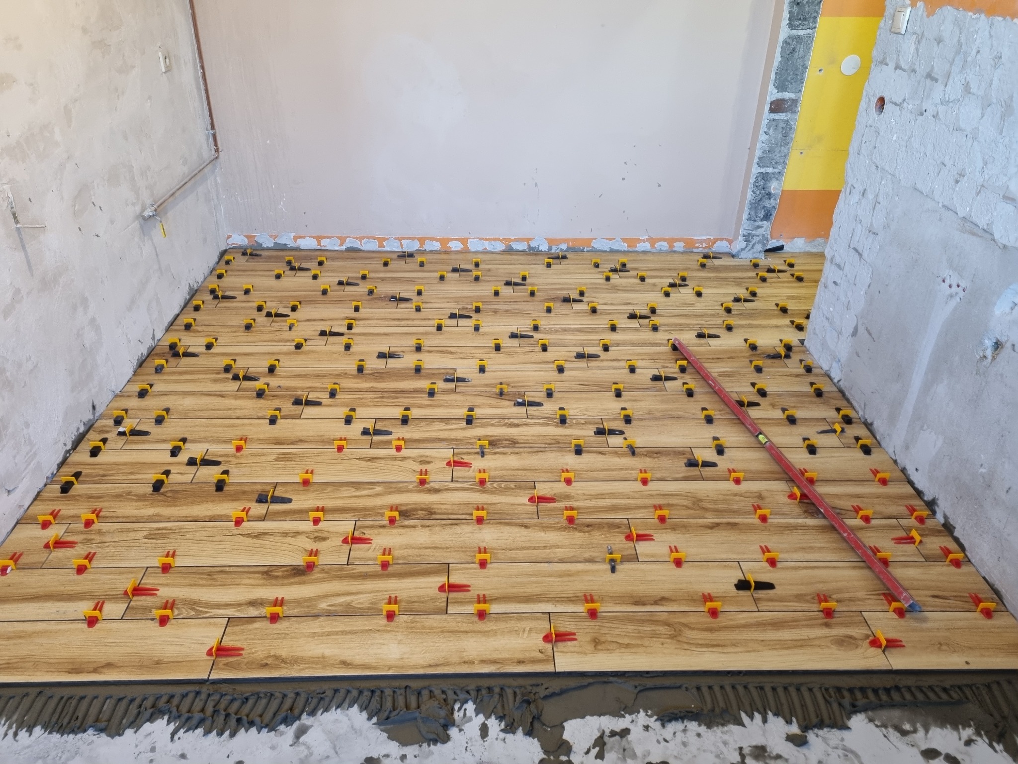 Układanie drewnopodobnych płytek podłogowych z użyciem systemu poziomowania, widoczna poziomica i surowa ściana w tle.