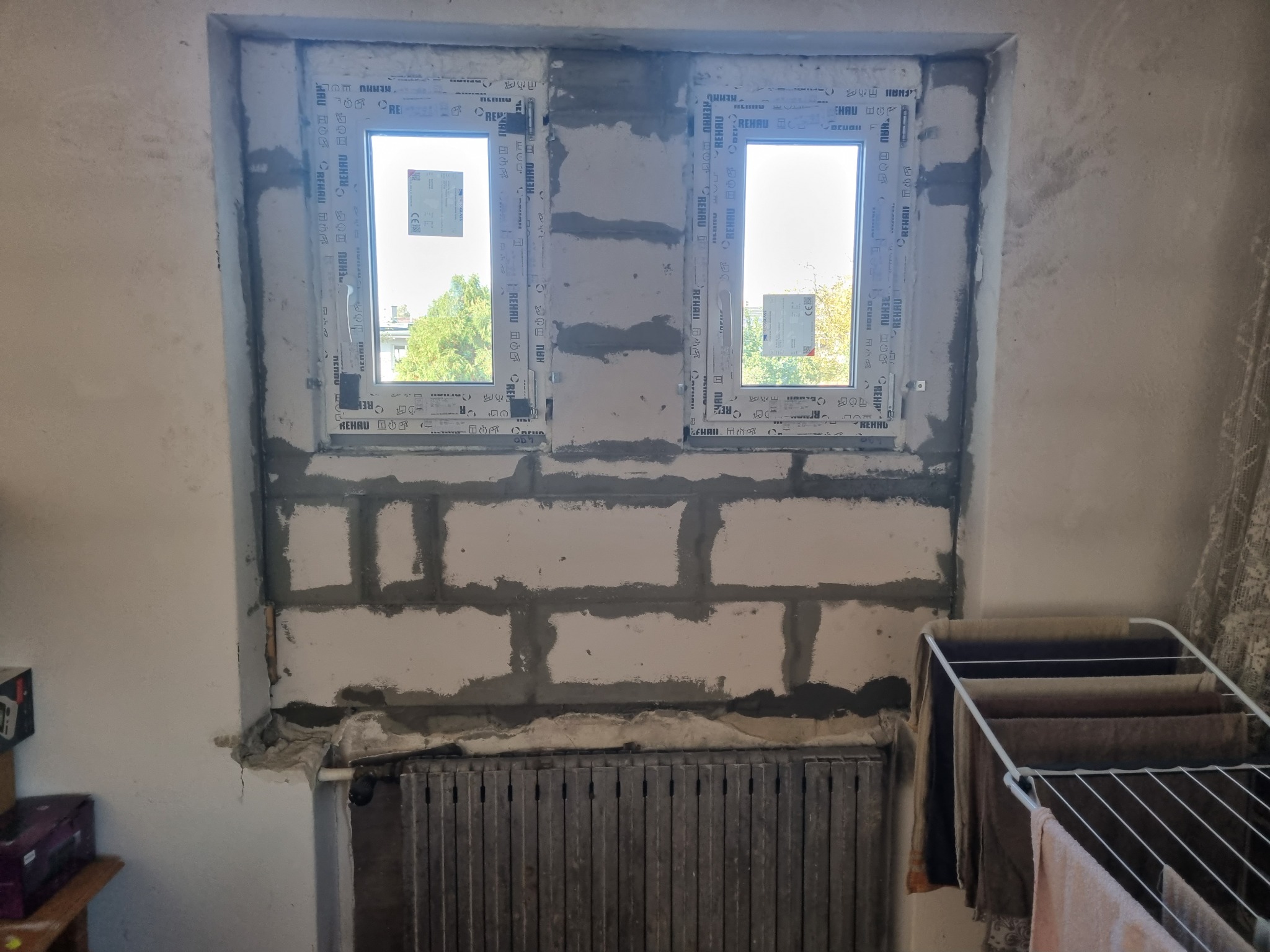 Dwa okna PCV zamontowane w surowej ścianie z betonu komórkowego, widoczne ślady zaprawy, grzejnik żeberkowy poniżej, suszarka z praniem po prawej stronie.
