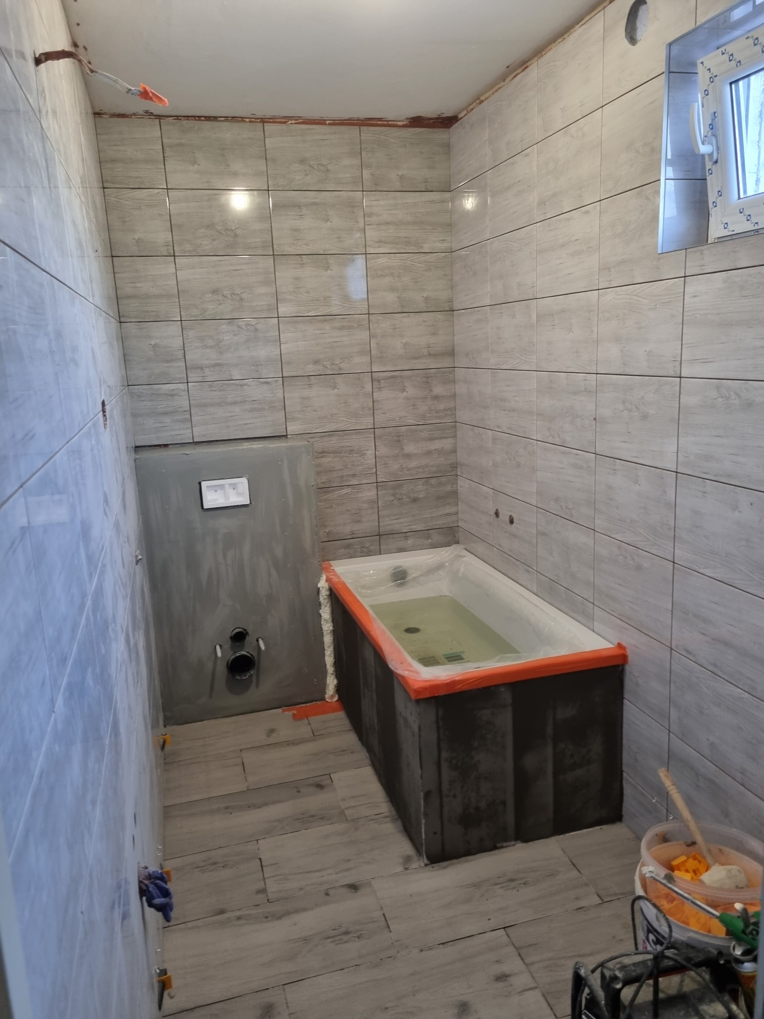 Łazienka w trakcie remontu, widoczne płytki imitujące drewno na ścianach i podłodze, zabudowana wanna zabezpieczona folią, oraz elementy instalacji podtynkowych. Na podłodze widoczne narzędzia...