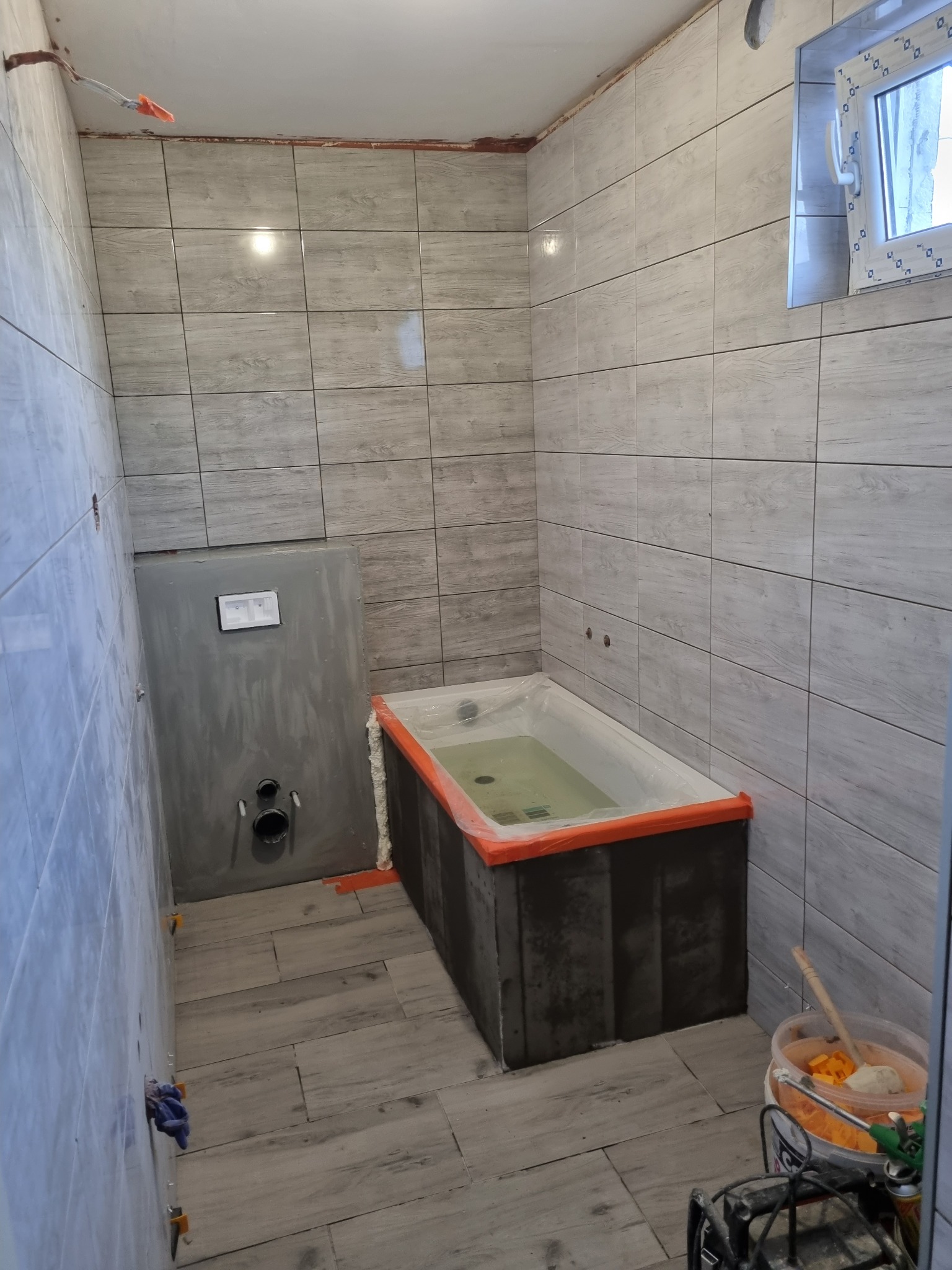 Wnętrze łazienki w trakcie remontu: wyłożone płytkami ściany i podłoga imitujące drewno, zabudowana wanna zabezpieczona folią, częściowo obudowany stelaż WC, widoczny pojemnik z zaprawą i narzędzia...