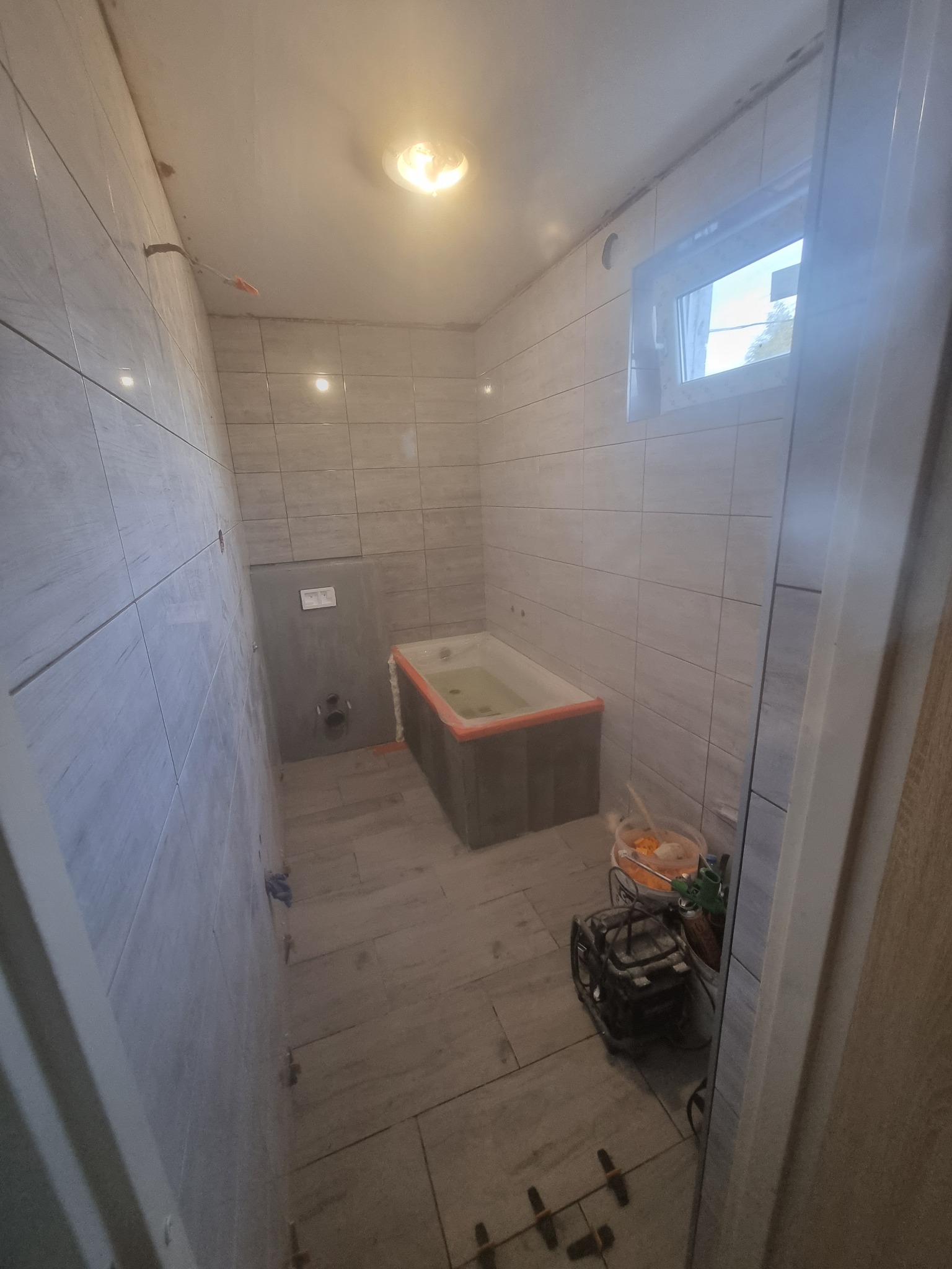 Wnętrze łazienki w trakcie remontu: świeżo położone szare płytki na ścianach i podłodze, wanna obudowana płytami, widoczne narzędzia i wiadro z zaprawą, okno z widokiem na zewnątrz.