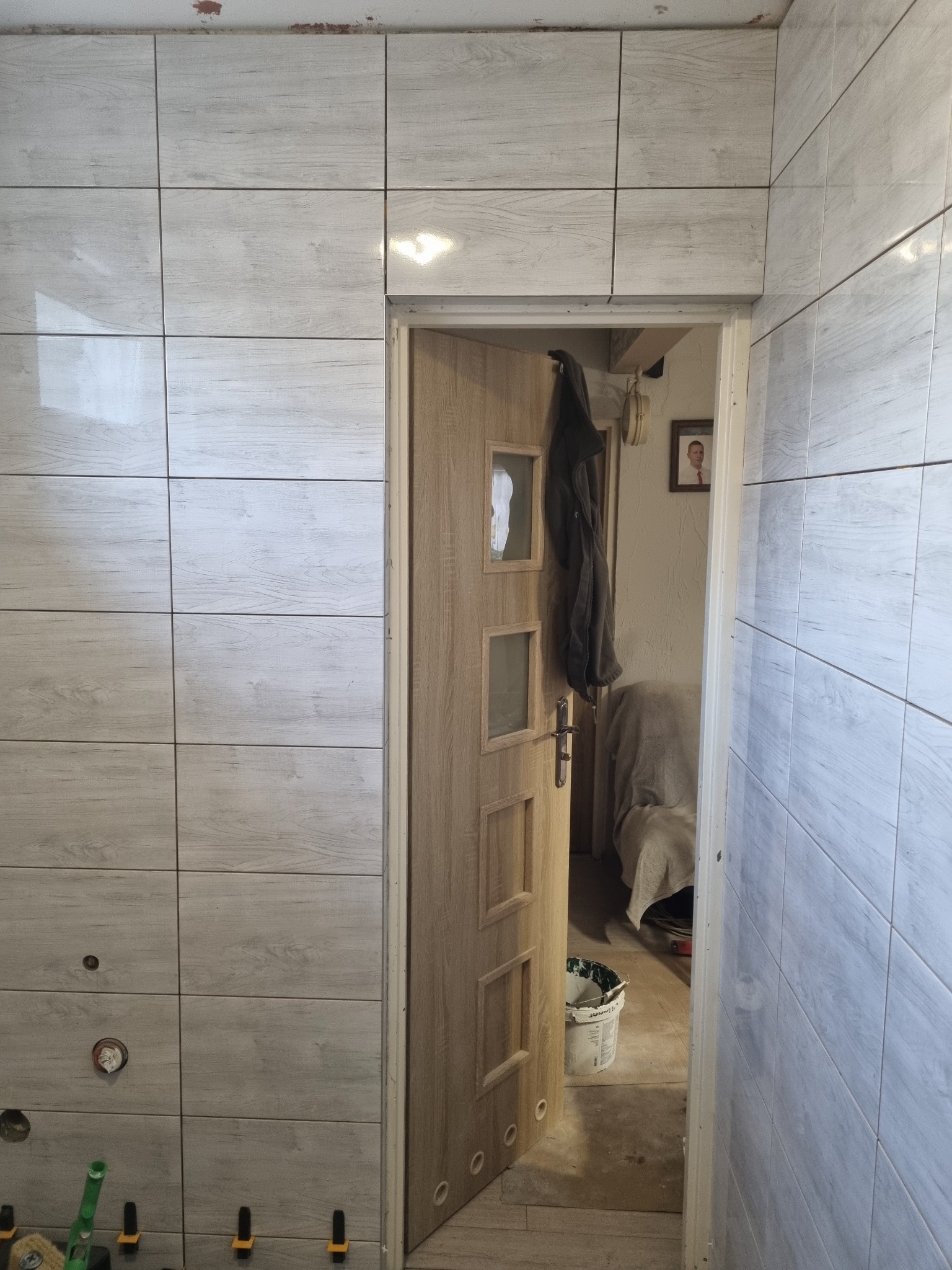Świeżo położone, jasne płytki imitujące drewno na ścianie w łazience w Siedlcu, widoczne narzędzia glazurnicze i otwarte drzwi do wnętrza.