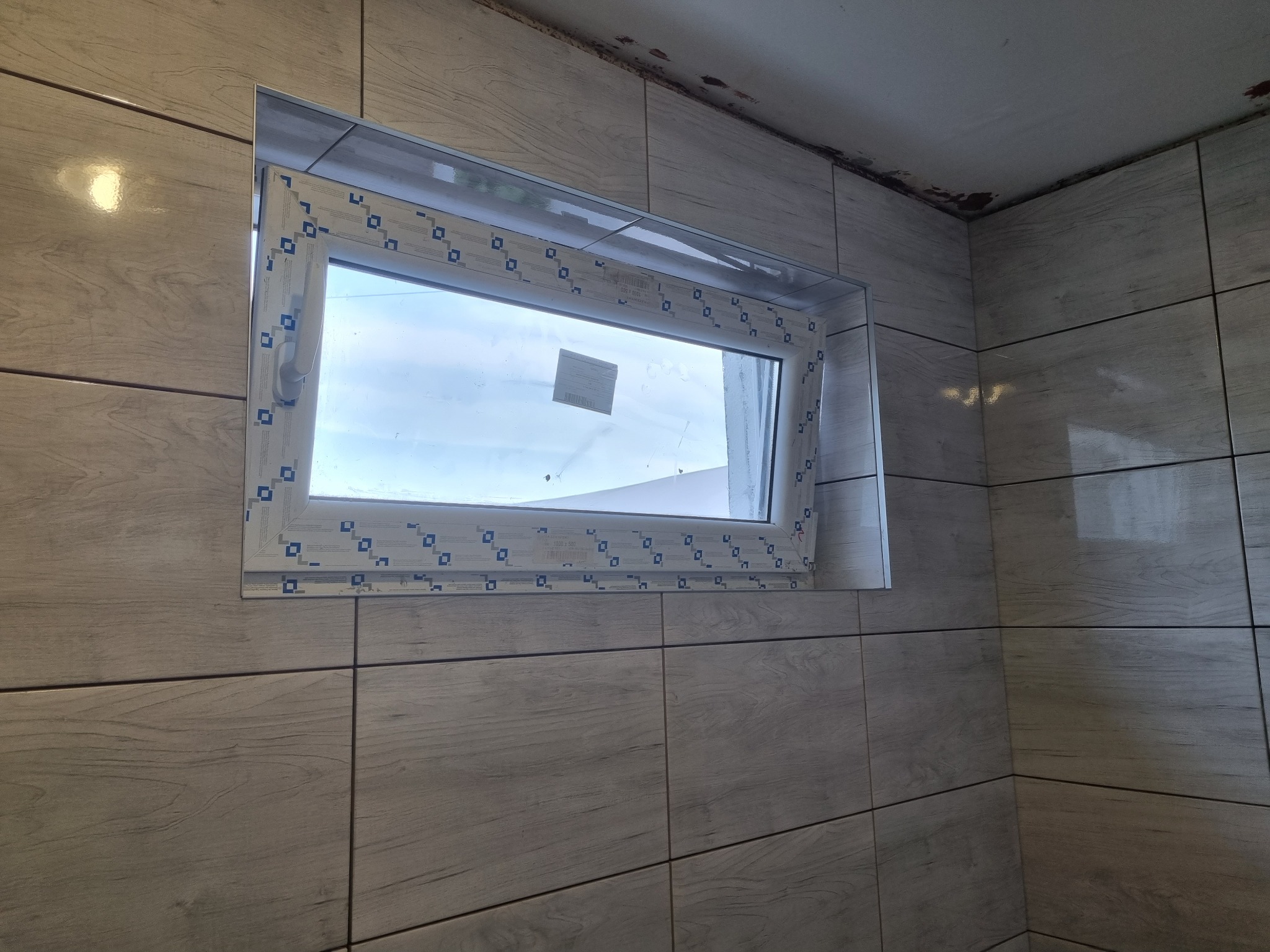 Okna PCV osadzone w ścianie wyłożonej płytkami imitującymi drewno, widoczny fragment sufitu z niewielkimi plamami.