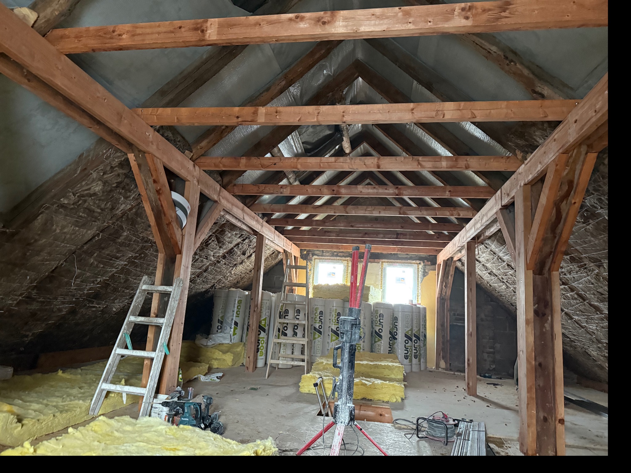 Poddasze w trakcie adaptacji: drewniana konstrukcja, izolacja z wełny mineralnej, drabina i narzędzia. Widoczne okna i surowy stan ścian.