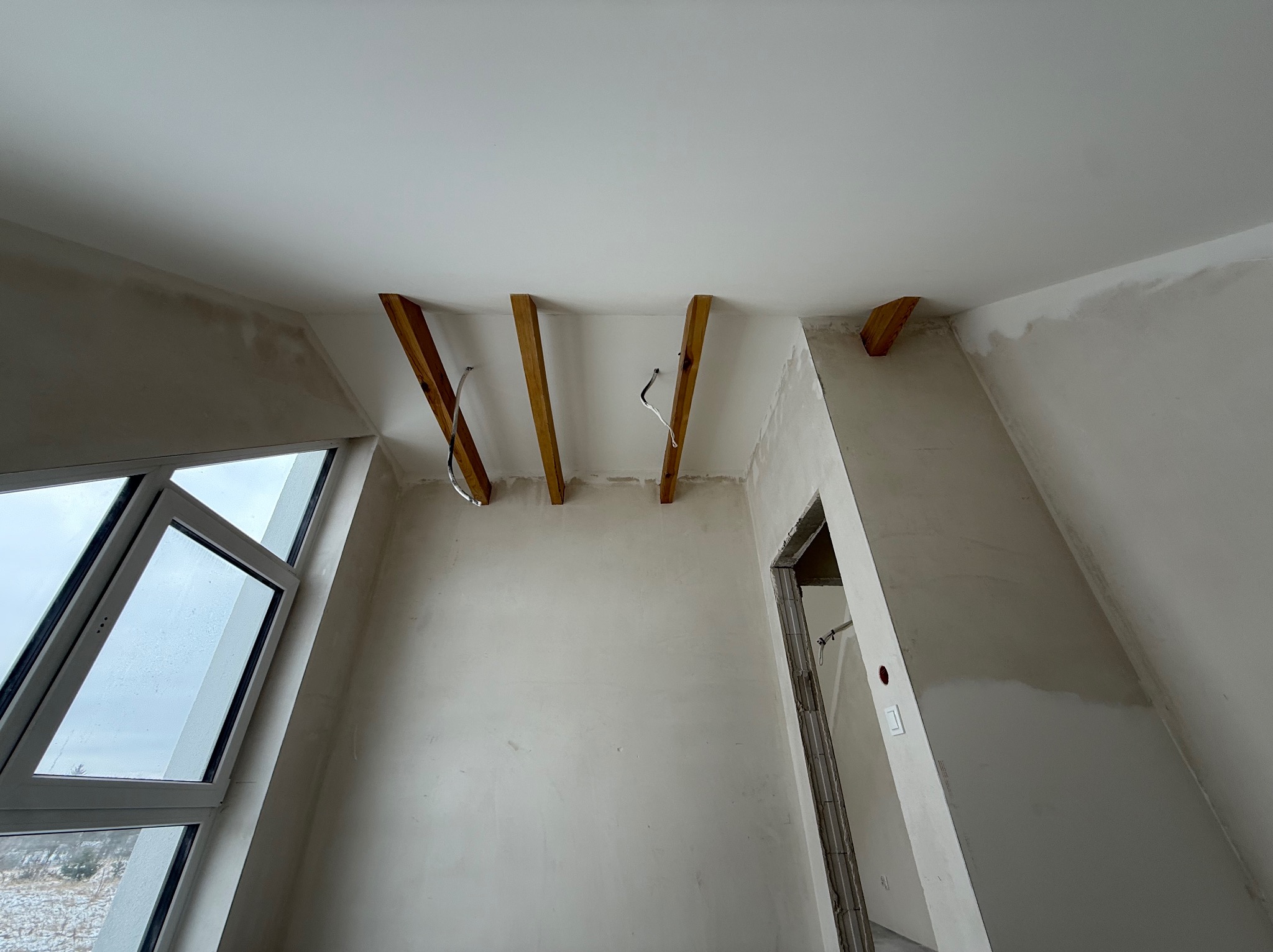 Surowe wnętrze z drewnianymi belkami pod sufitem i oknem. Ściany przygotowane do dalszych prac wykończeniowych, widoczne ślady szpachlowania. Stan surowy.