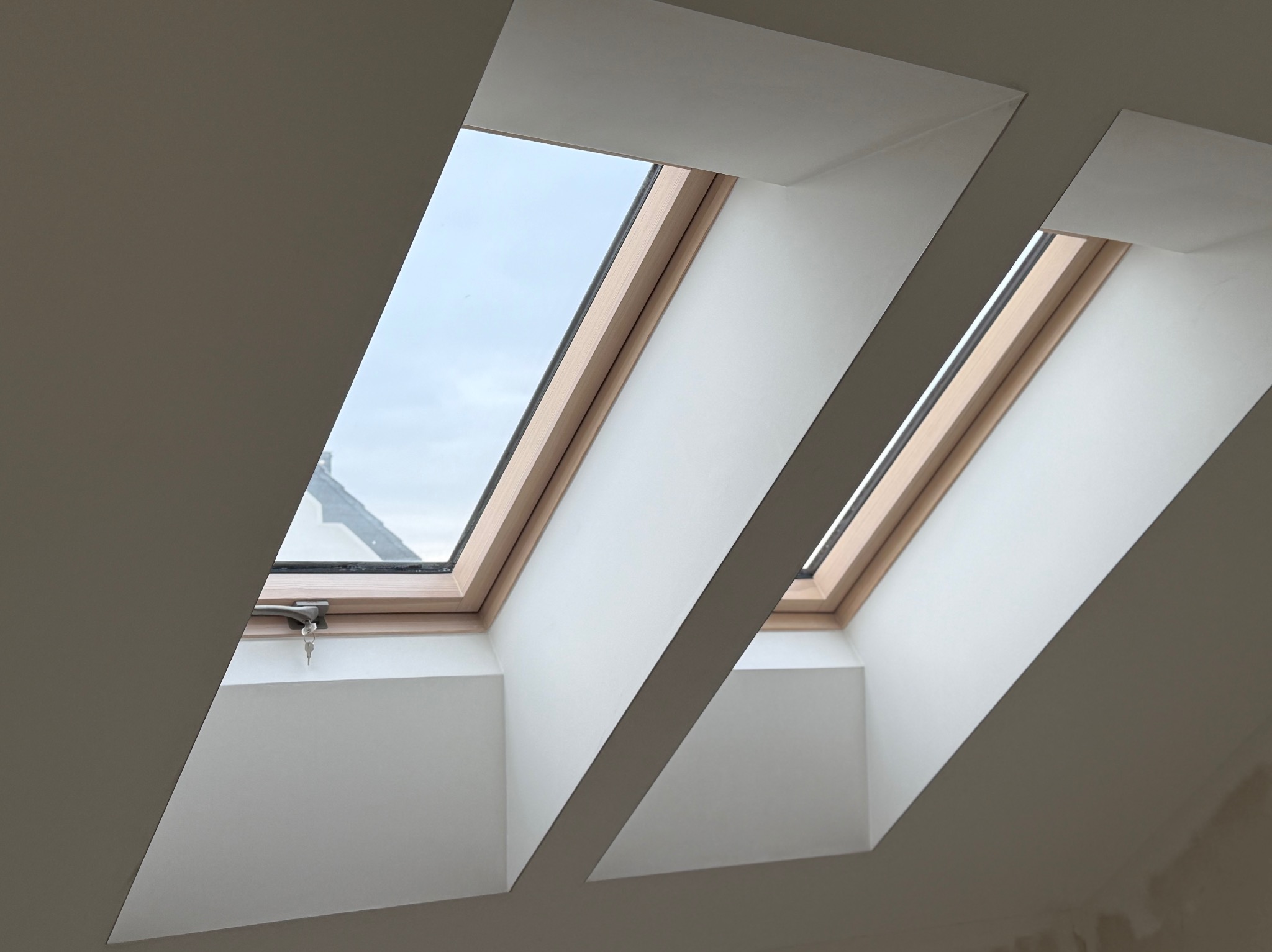 Dwa okna dachowe z drewnianą ramą, w białej wnęce, z kluczykiem w zamku jednego z okien. Widok na niebo i fragment dachu sąsiedniego budynku.
