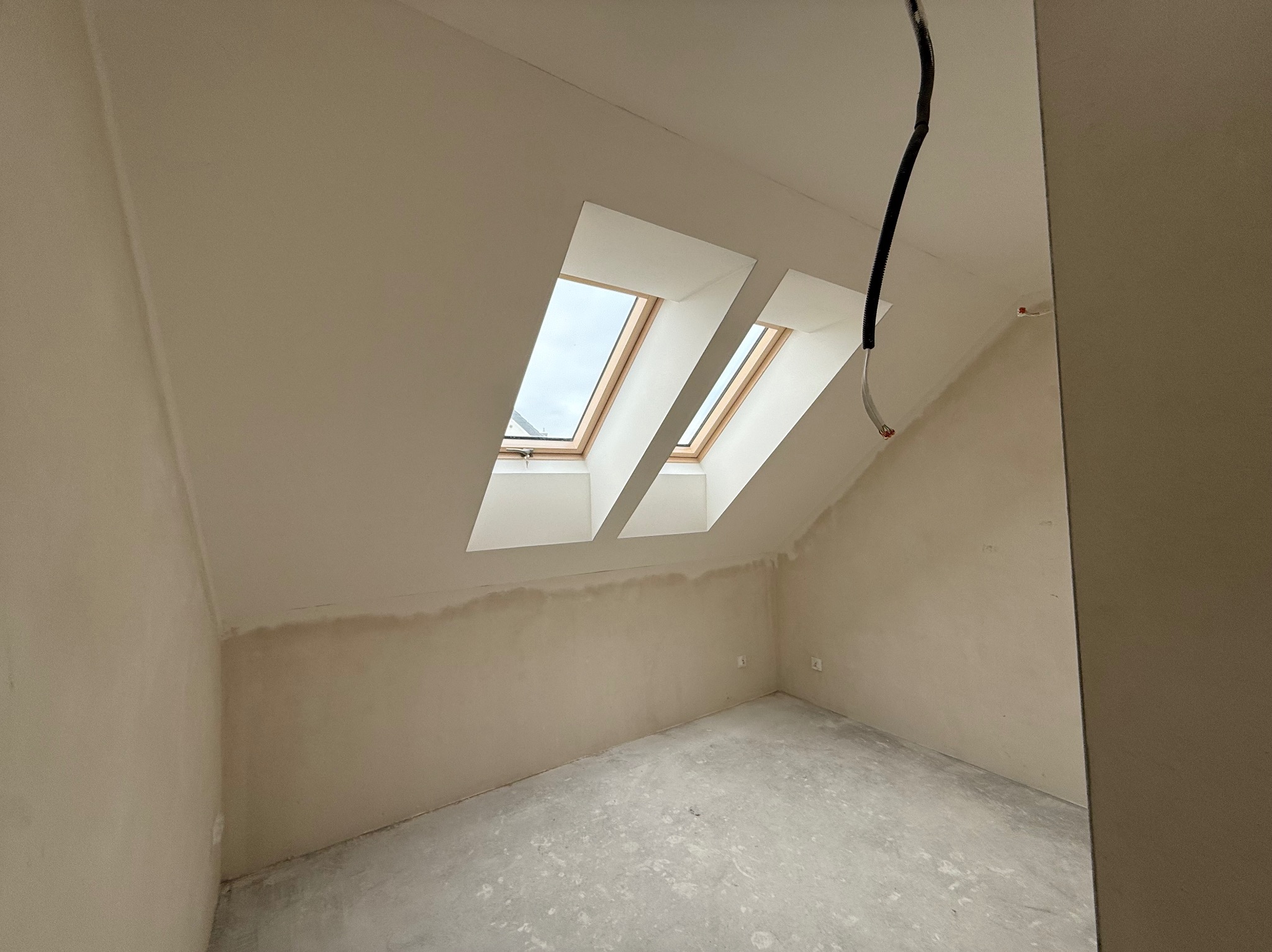 Pokój na poddaszu w trakcie wykańczania: dwa okna dachowe, ściany przygotowane do malowania, widoczne przewody elektryczne. Surowy stan wykończenia.