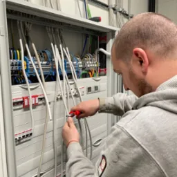 Elektryk w szarej bluzie obcina przewód w rozdzielnicy elektrycznej za pomocą czerwonych obcinaczek, widoczne liczne kable i zabezpieczenia.