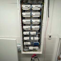 SuperElektroVolt - Otwarta skrzynka rozdzielcza z bezpiecznikami i przewodami w jasnym pomieszczeniu, widoczna drabina i fragment dłoni w dolnej części kadru.